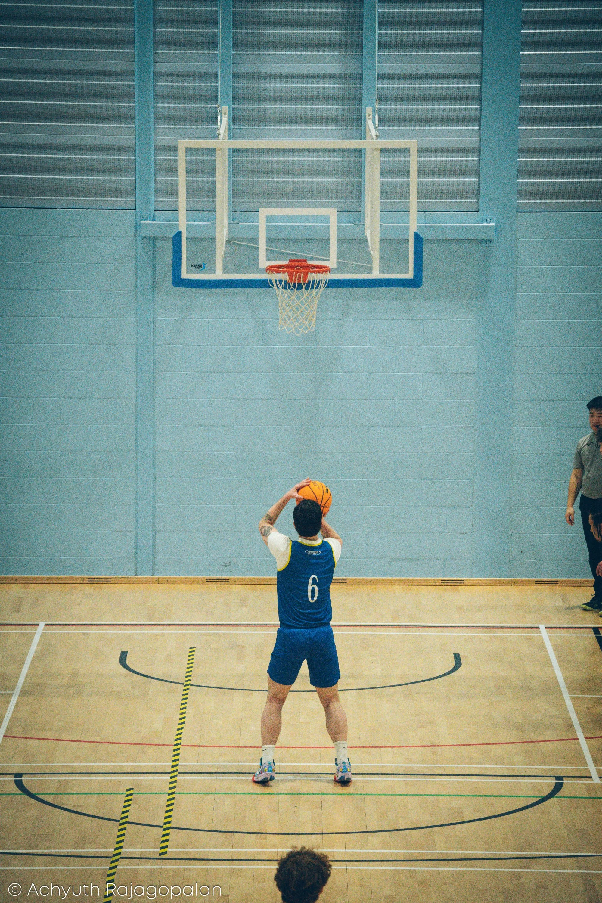 A person wearing a blue basketball uniform with the number 6 on the back, holding a basketball above their head on a basketball court, with part of another person on the right and a person with curly hair at the bottom. The background features a bask