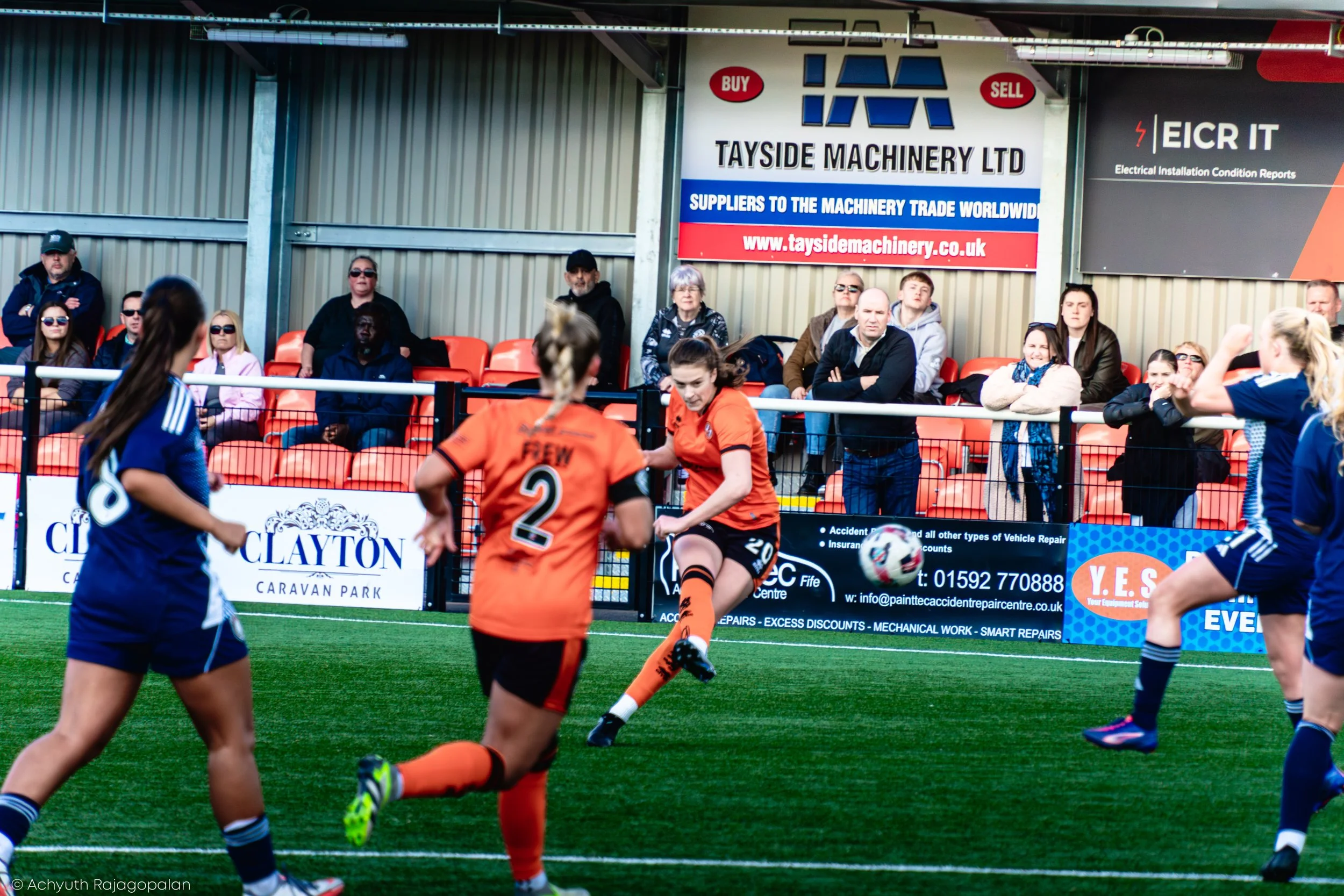 Women and girls playing soccer on a field, with spectators watching from bleachers in the background.