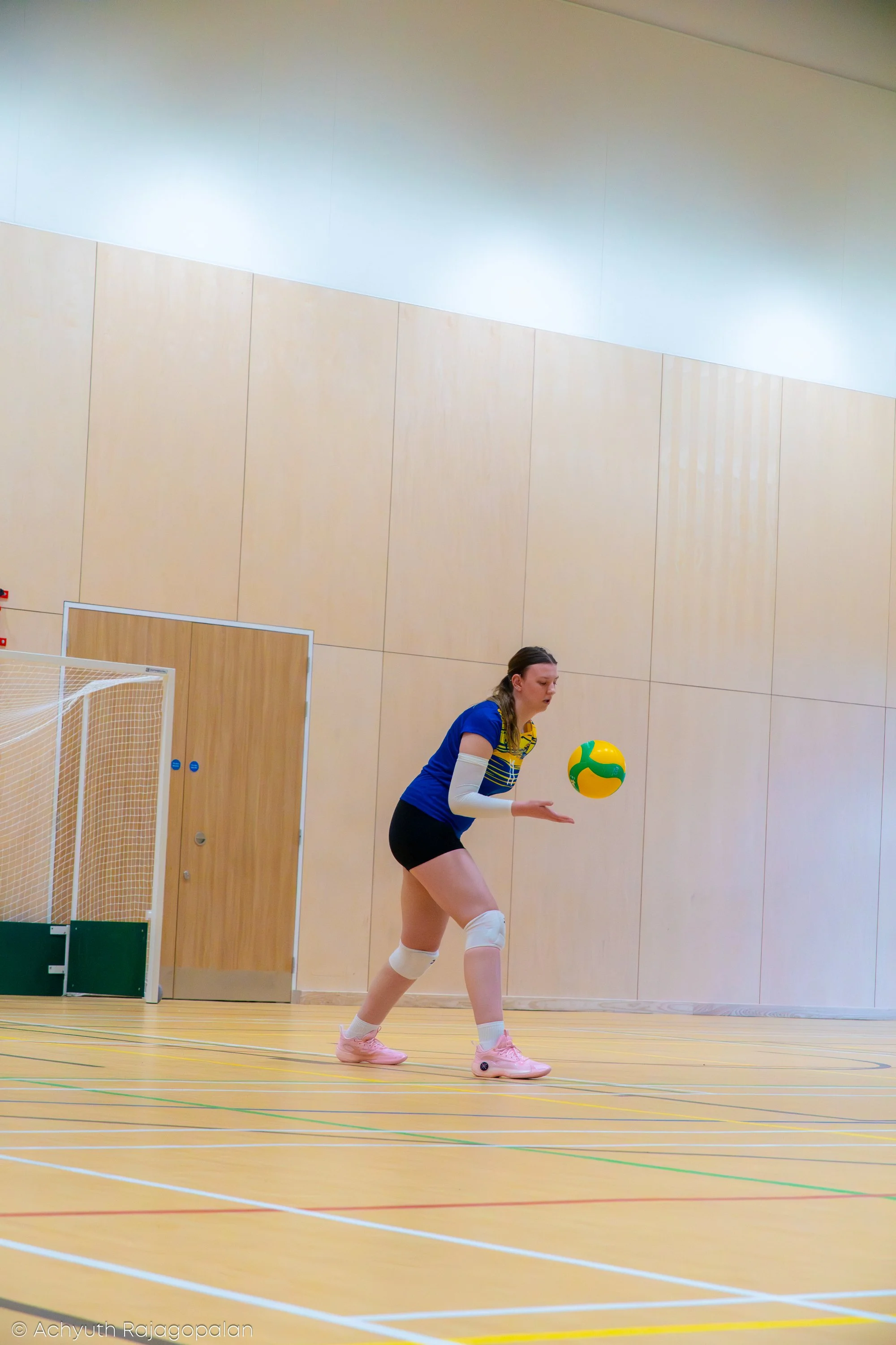 A female volleyball player in a blue sports shirt, black shorts, and pink shoes is preparing to serve or pass a volleyball on an indoor court with a wooden floor, beige walls, and a goalpost.