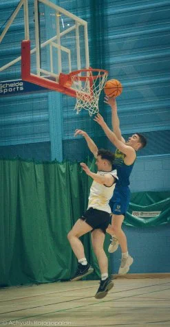 Two boys playing basketball indoors, one attempting a shot at the hoop while the other defends.