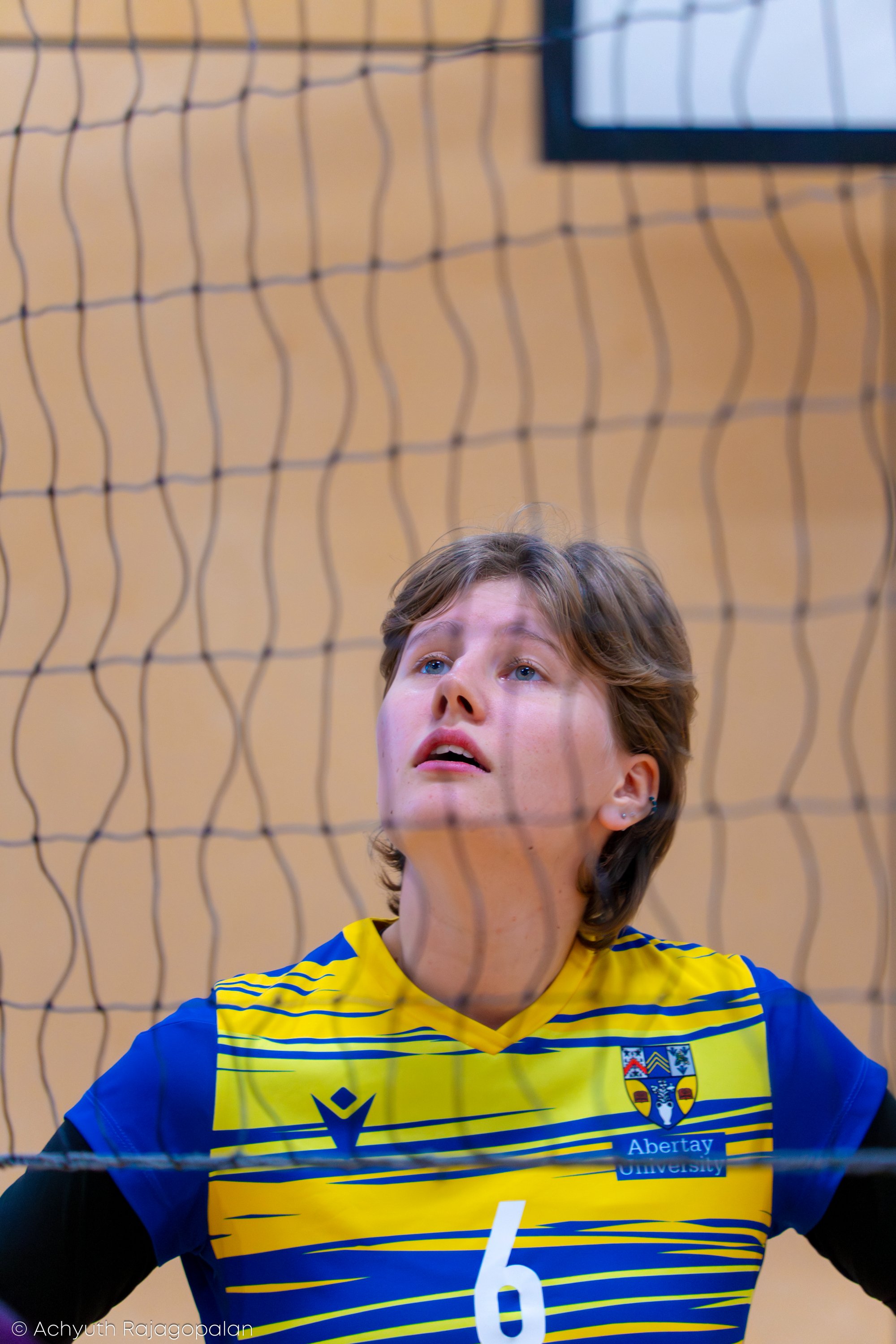 A woman with short brown hair and blue eyes is in front of a volleyball net, wearing a blue and yellow sports jersey with the logo of Abertay University. The photo is taken from a low angle, with part of a scoreboard visible in the background.
