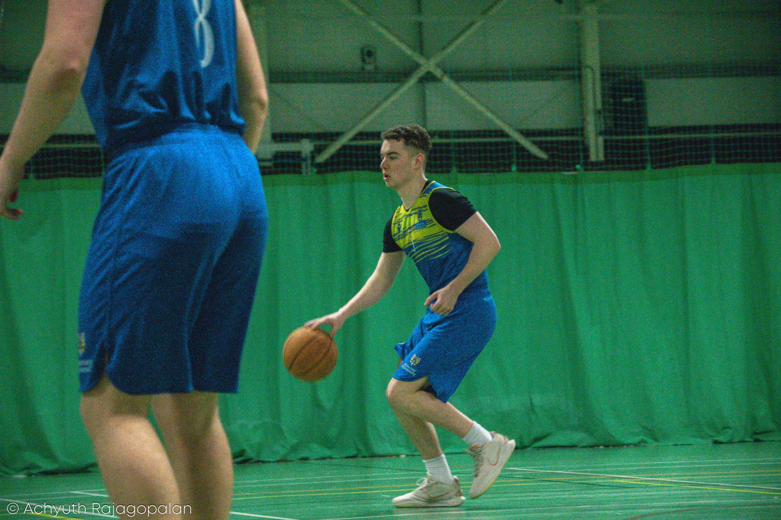 Two basketball players are practicing inside a gymnasium with green walls and a netted ceiling. One player, wearing a yellow and black jersey, is dribbling a basketball, while the other player, dressed in blue sportswear, stands nearby.