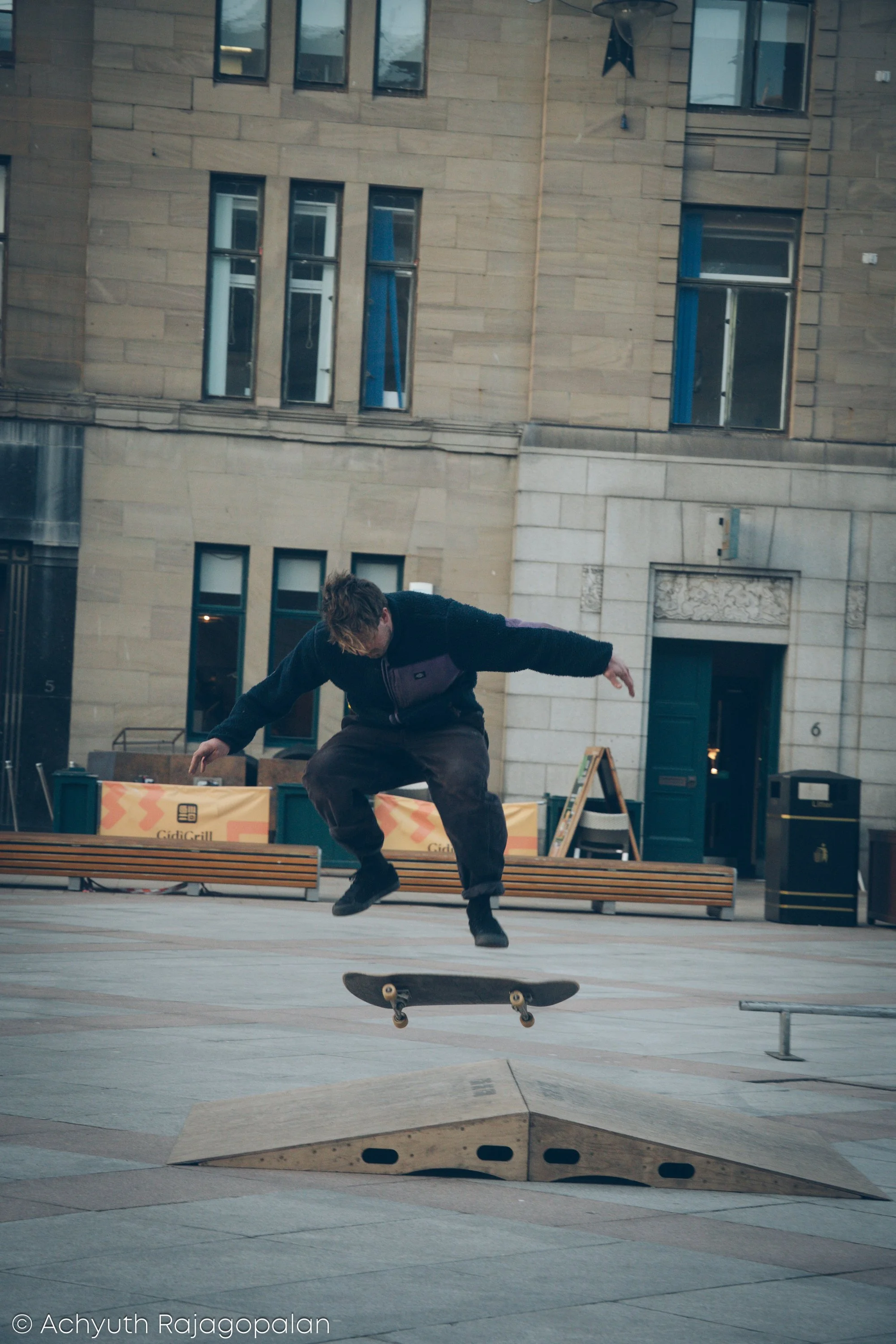 A skateboarder performing a trick in an urban setting with multi-story buildings in the background. He is mid-air above a wooden ramp.
