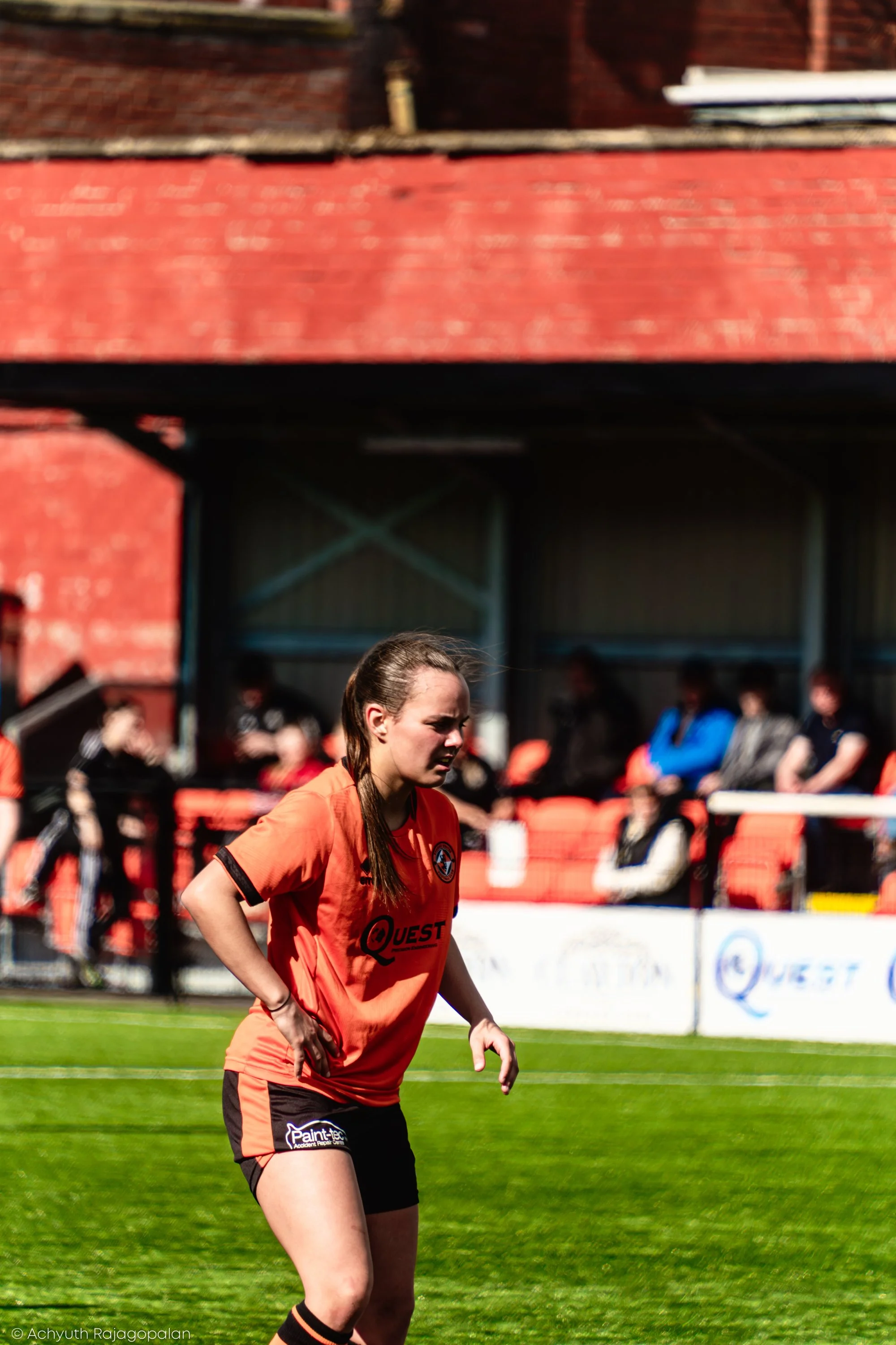 A female soccer player on the field during a game, wearing an orange jersey and black shorts, with a crowd and a brick building in the background.