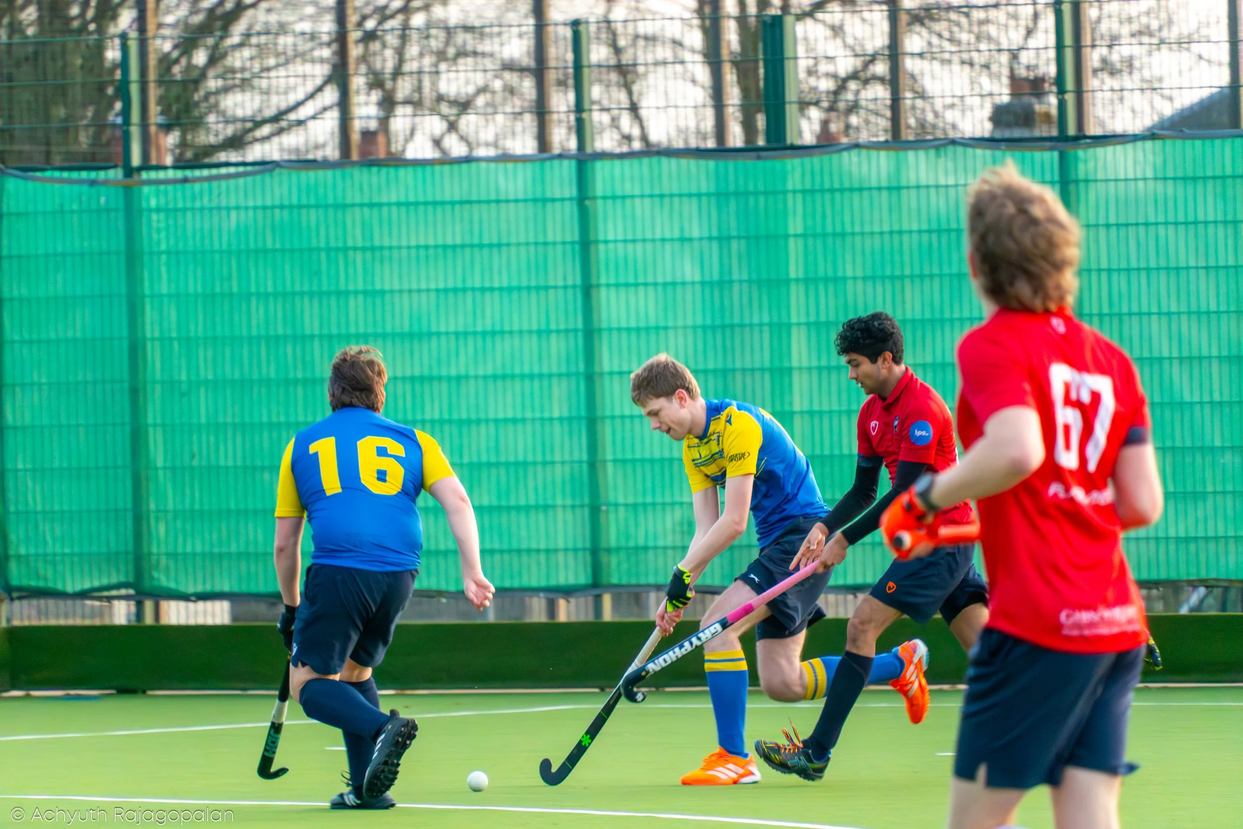 Young hockey players, boys and girls, in colorful jerseys and shorts, playing hockey on an outdoor field with a green fence and trees in the background.