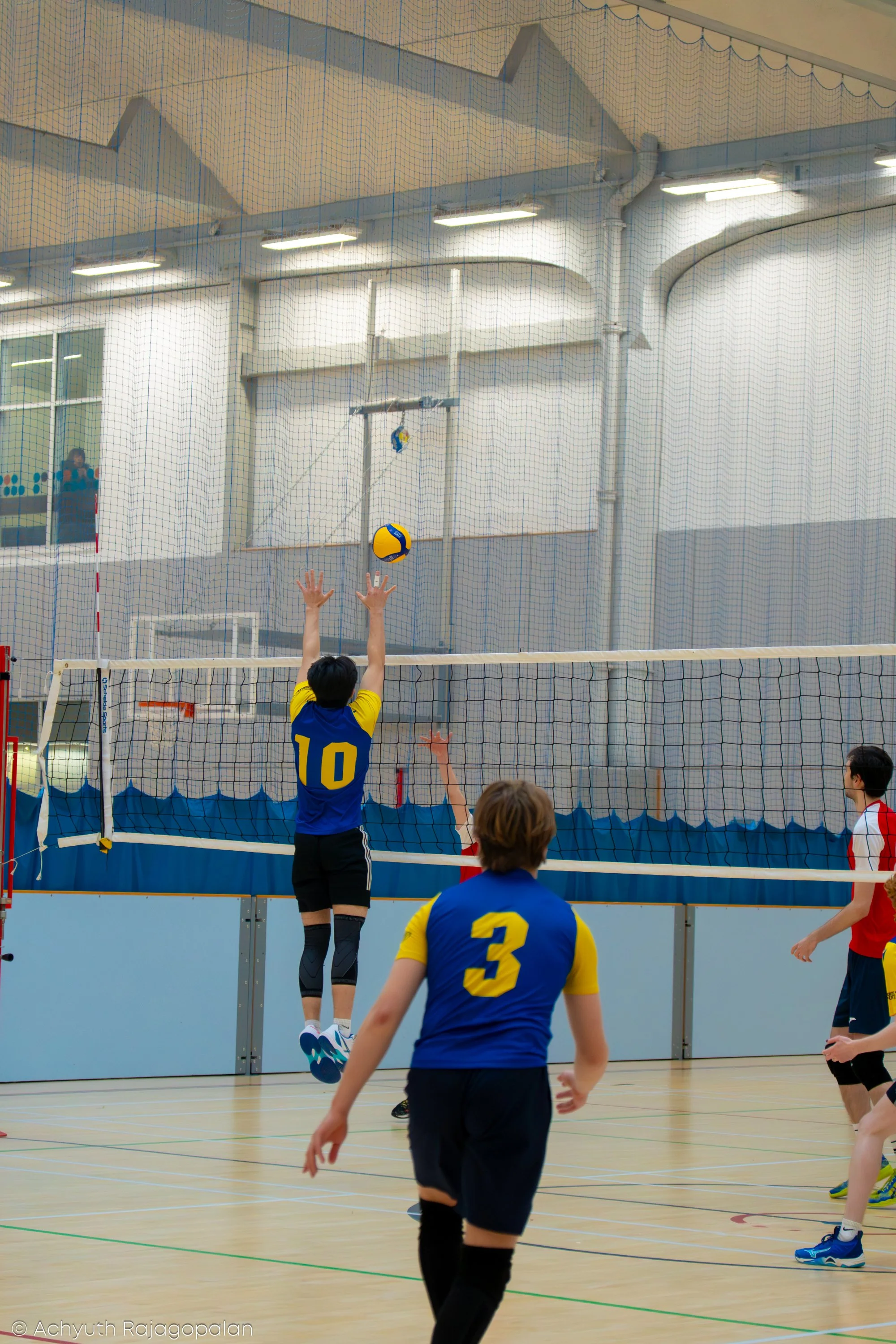 A volleyball game in progress at an indoor gym. A player in a blue and yellow uniform with the number 10 is jumping to hit the ball over the net, while another player in a blue and yellow uniform with the number 3 is preparing to receive or defend. T