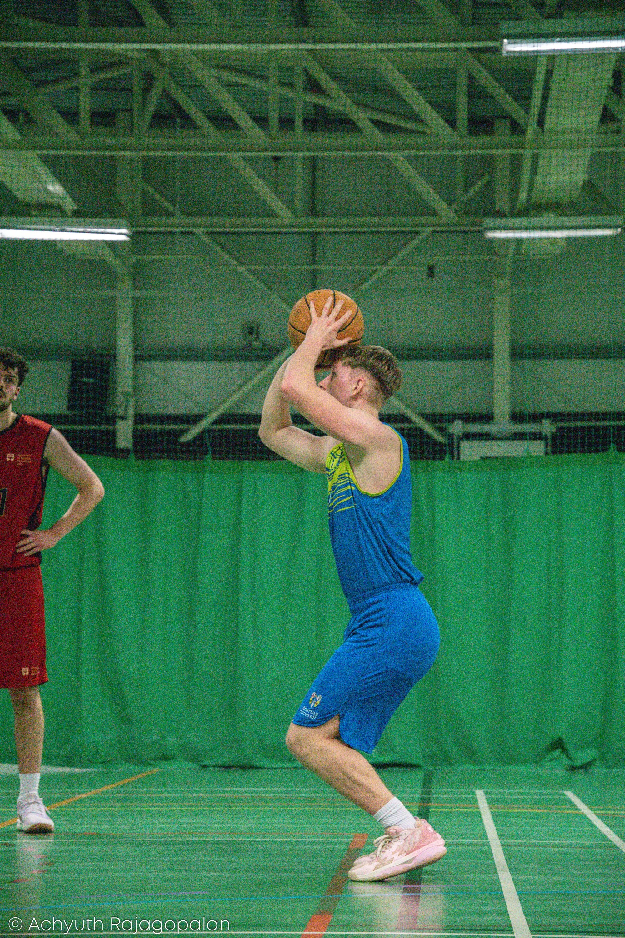 A young woman in a blue sports uniform is preparing to shoot a basketball on an indoor court. She is holding the basketball above her head with both hands, positioning for a shot. Another player in a red uniform stands nearby with hands on hips. The 