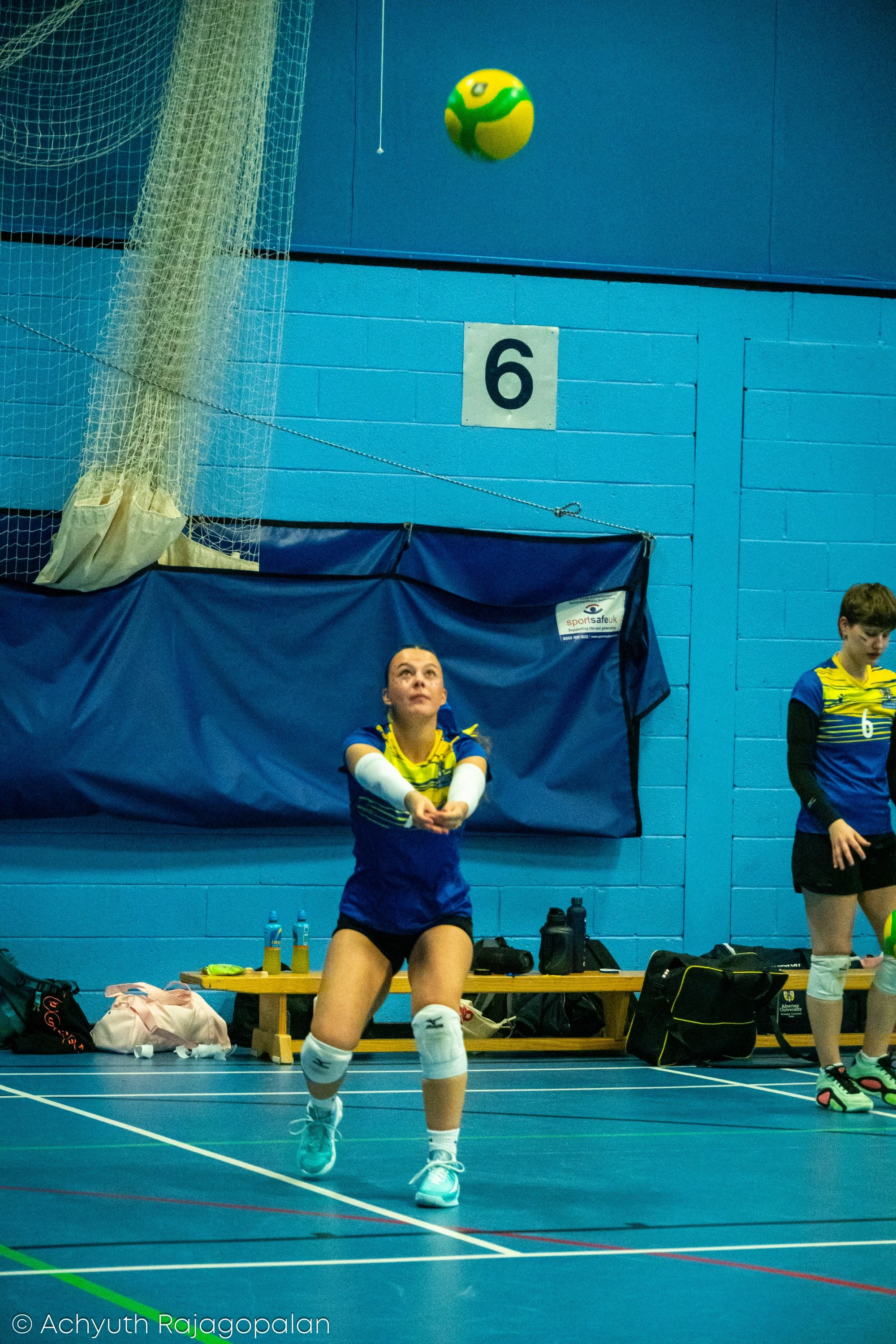 A female volleyball player in a blue and yellow uniform is seated on a bench in an indoor sports hall, preparing to serve the volleyball. The volleyball is in mid-air above her. There are some water bottles, bags, and equipment behind her.