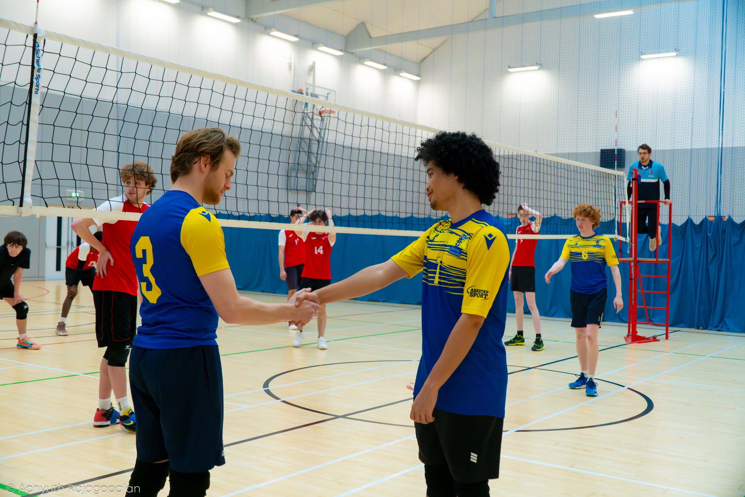 Two volleyball players from opposing teams shaking hands at the net after a match in an indoor gym.