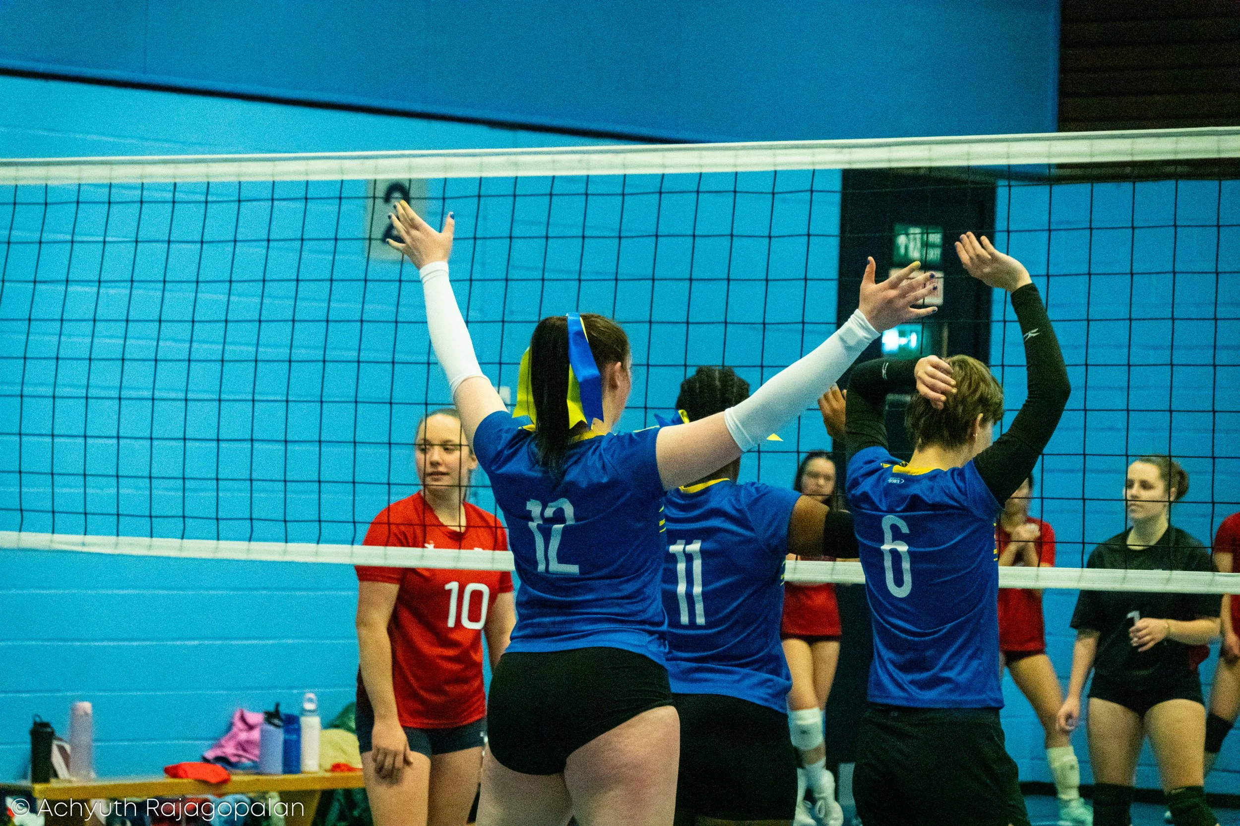A volleyball game in progress with players near the net, some in blue uniforms and others in red uniforms, inside a gymnasium.