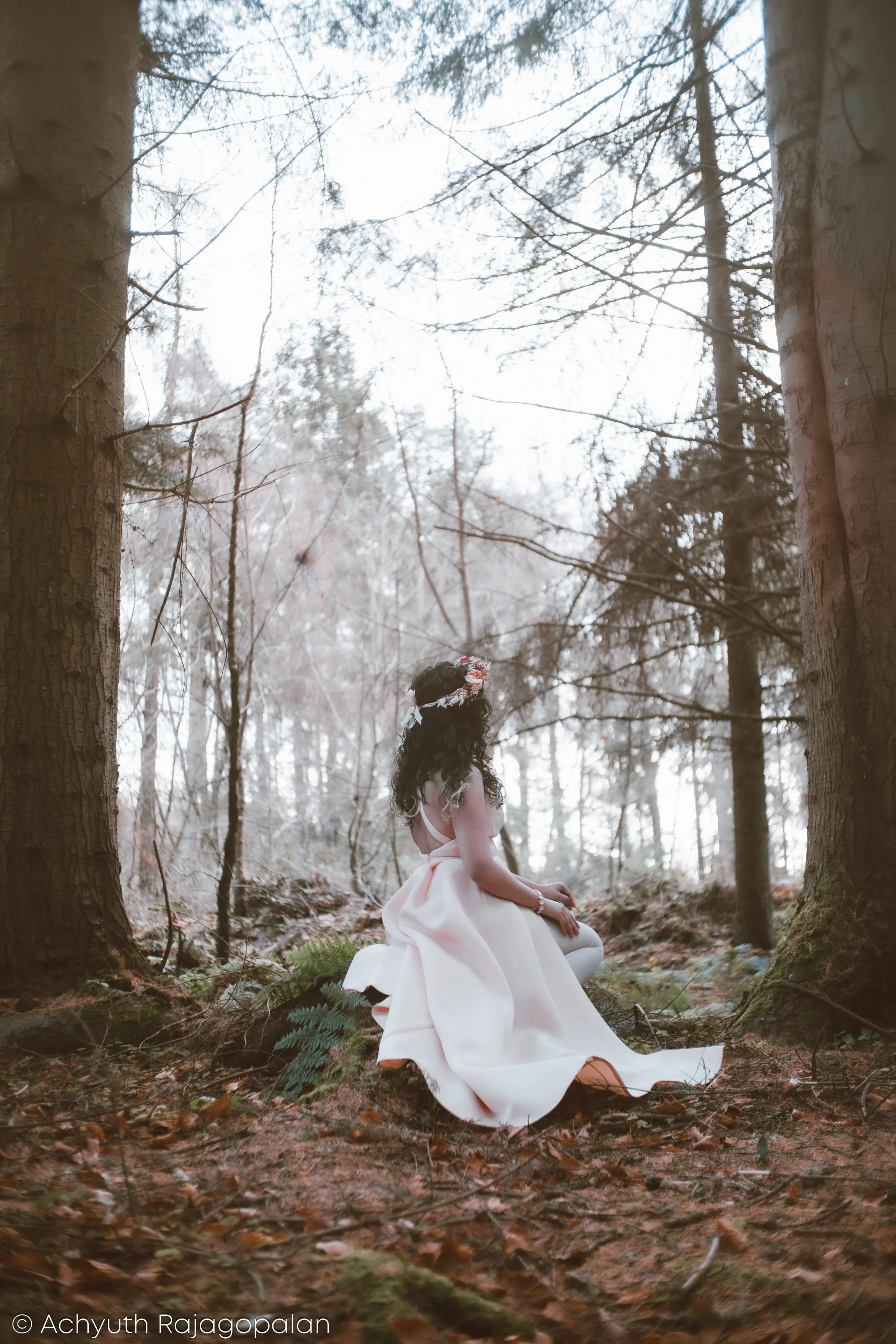 A woman with dark, curly hair wearing a light pink or white dress with a bow, sitting in a forest surrounded by trees, moss, and fallen leaves, with a cloudy sky overhead.