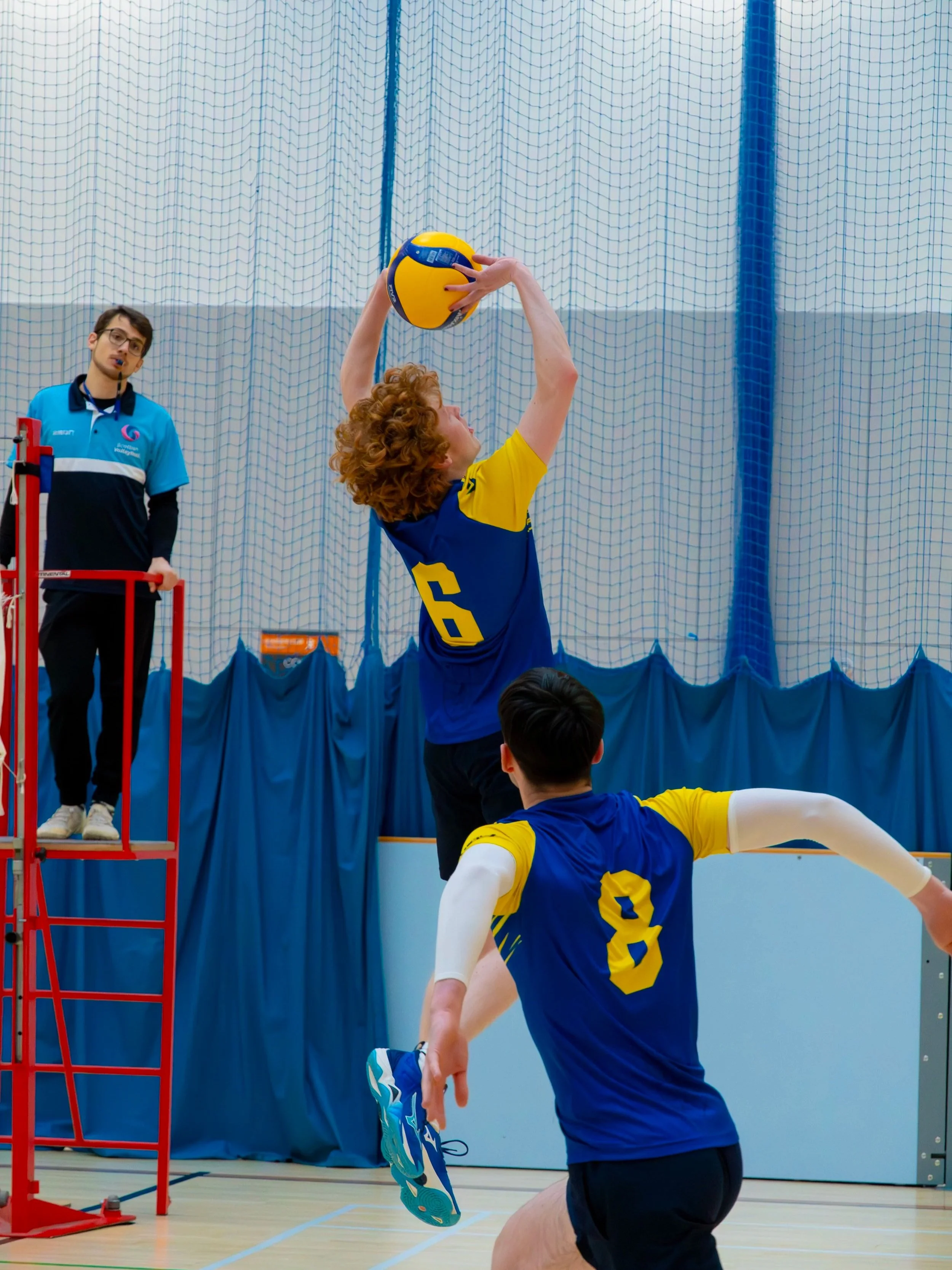 A volleyball game in progress with a player jumping to hit the ball over the net, an official watching attentively, and a person lying on the ground, wearing blue jerseys with yellow accents, indoors with a blue netted wall in the background.
