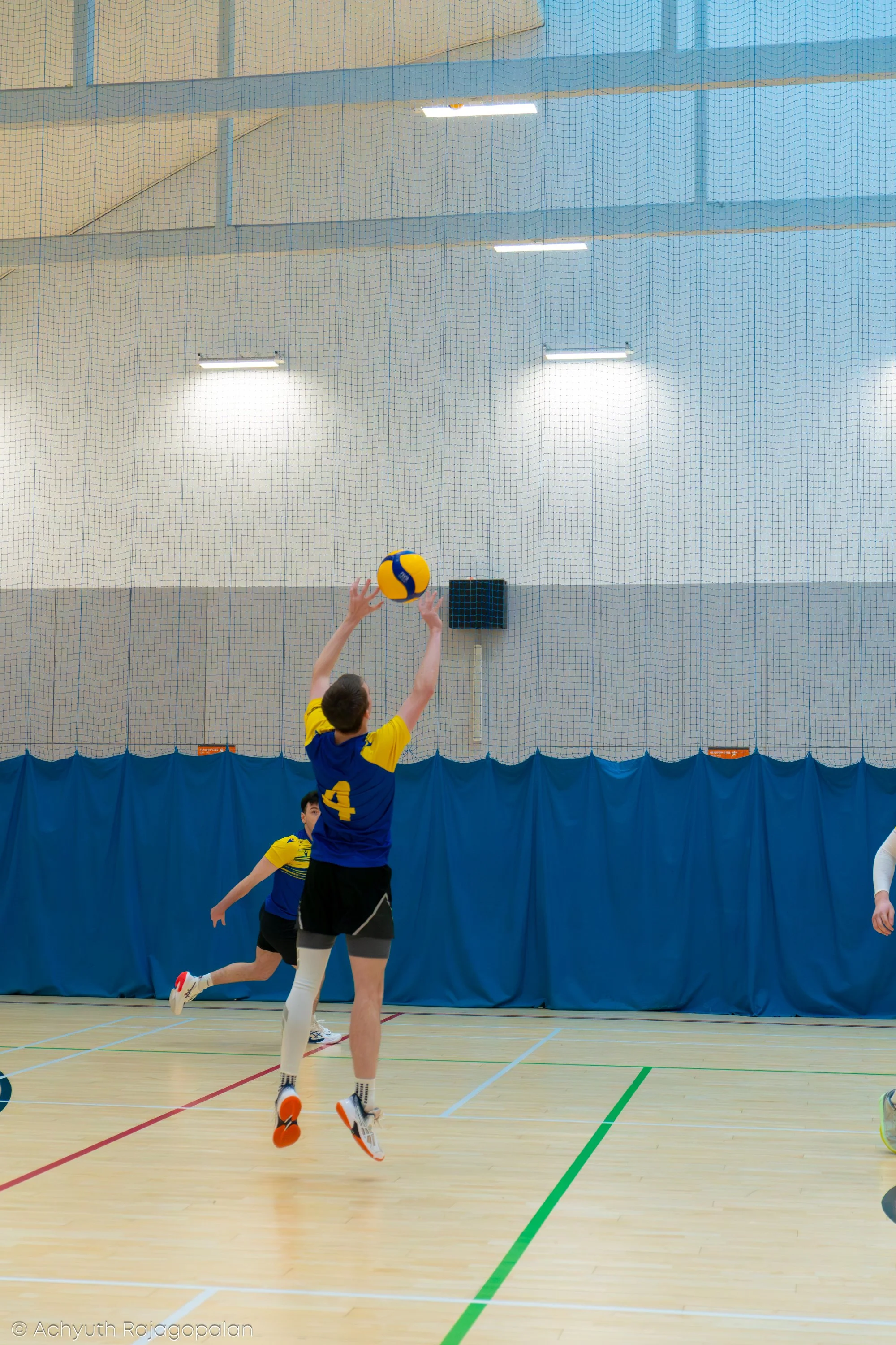 A volleyball game in progress with players on an indoor court, some jumping and reaching for the ball, with a gymnasium backdrop and nets.