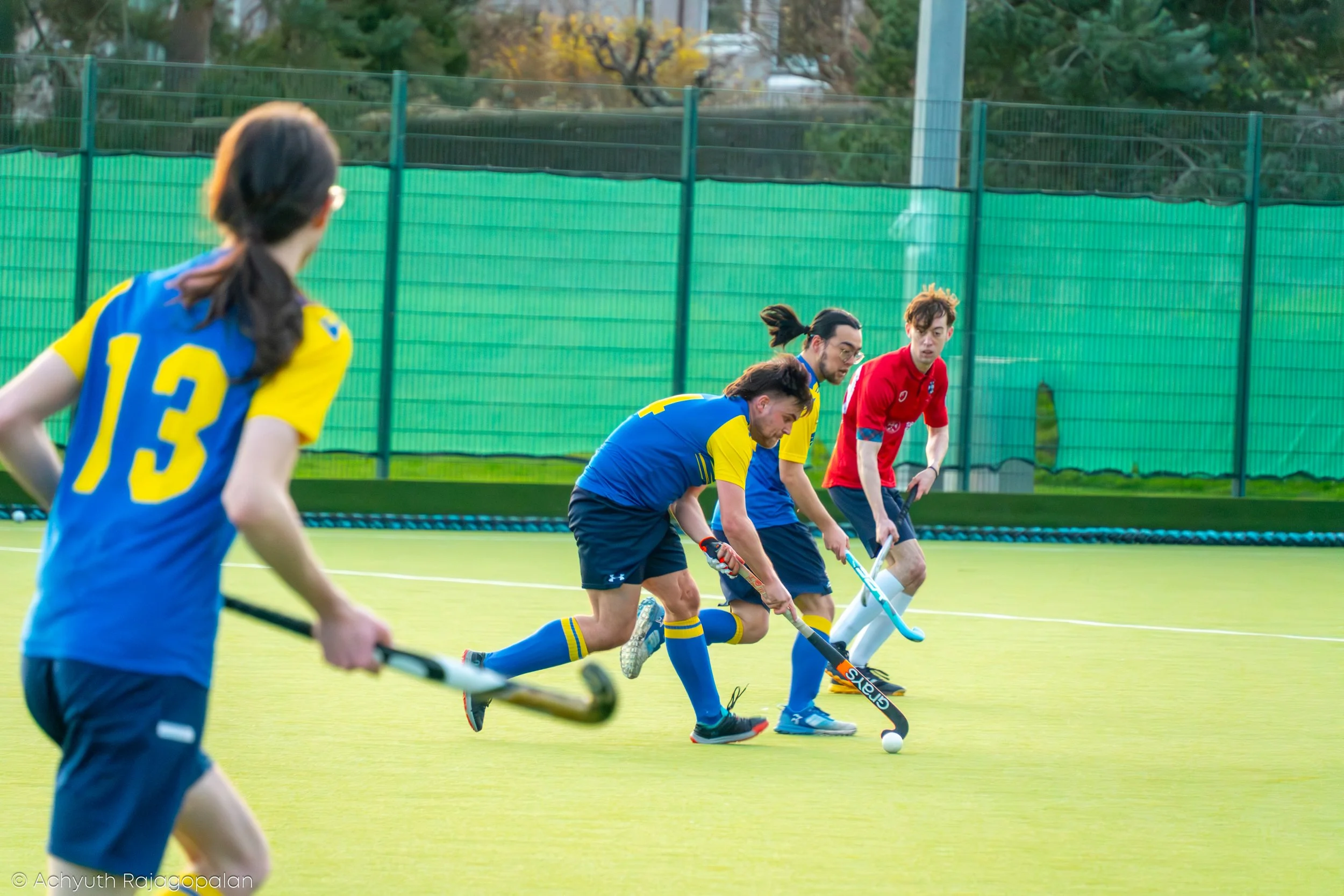 A group of players in blue and yellow and one in red uniforms playing field hockey on an outdoor field with green fencing and trees in the background.