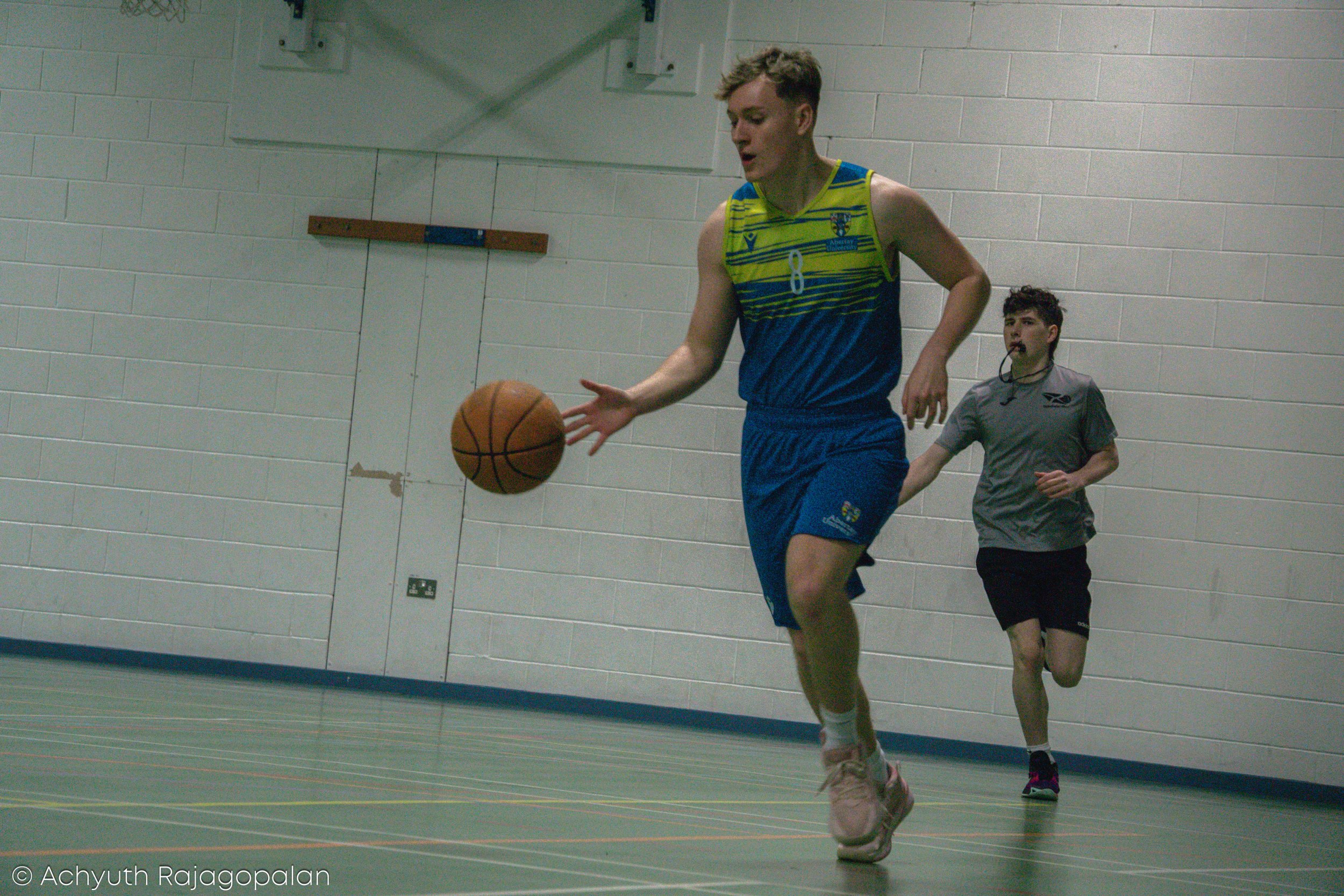 A young male athlete dribbling a basketball indoors, wearing a blue and yellow sports uniform with the number 8, with a referee or coach following in the background.