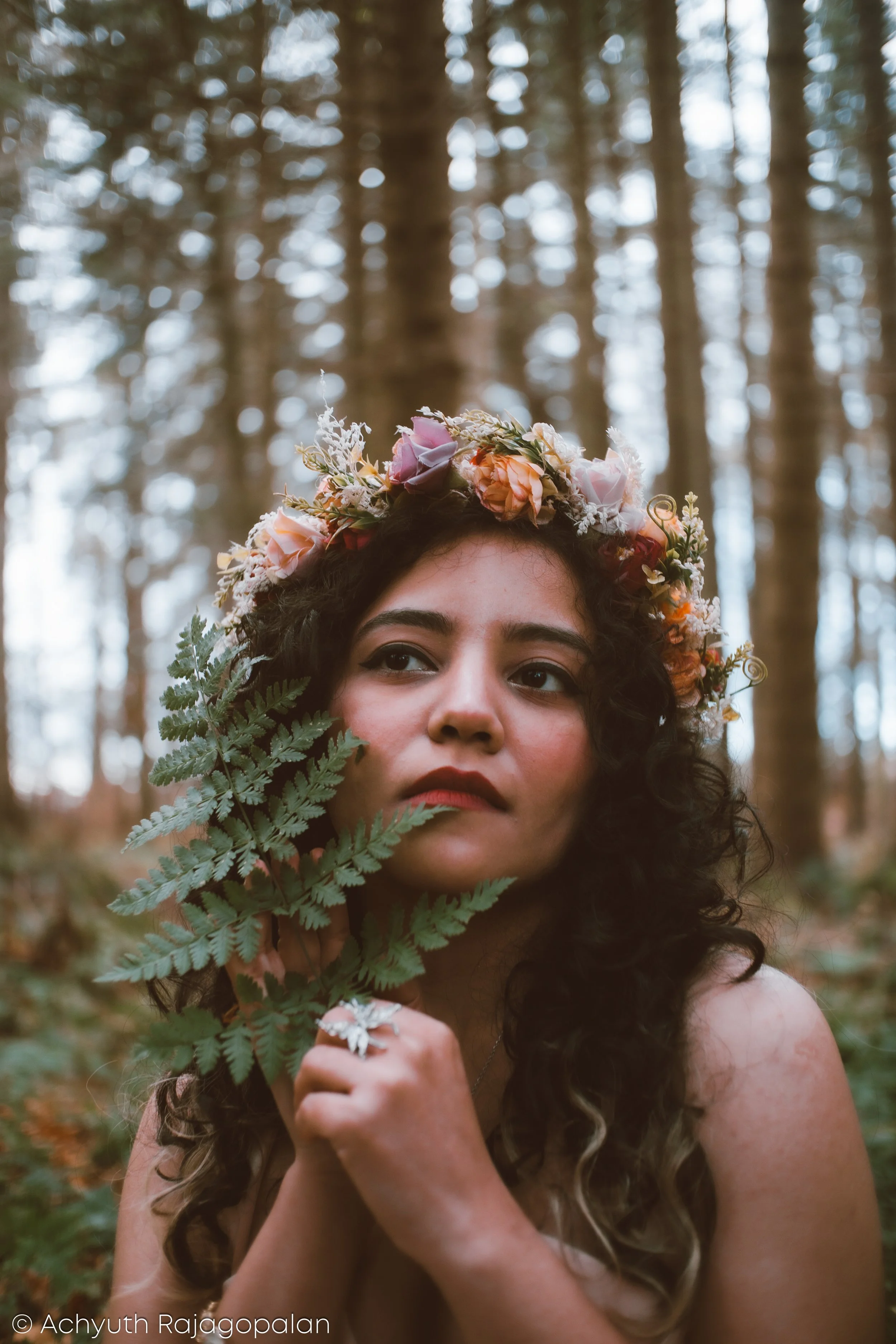 A woman with dark curly hair wearing a floral crown in a forest, holding a green fern leaf near her face.