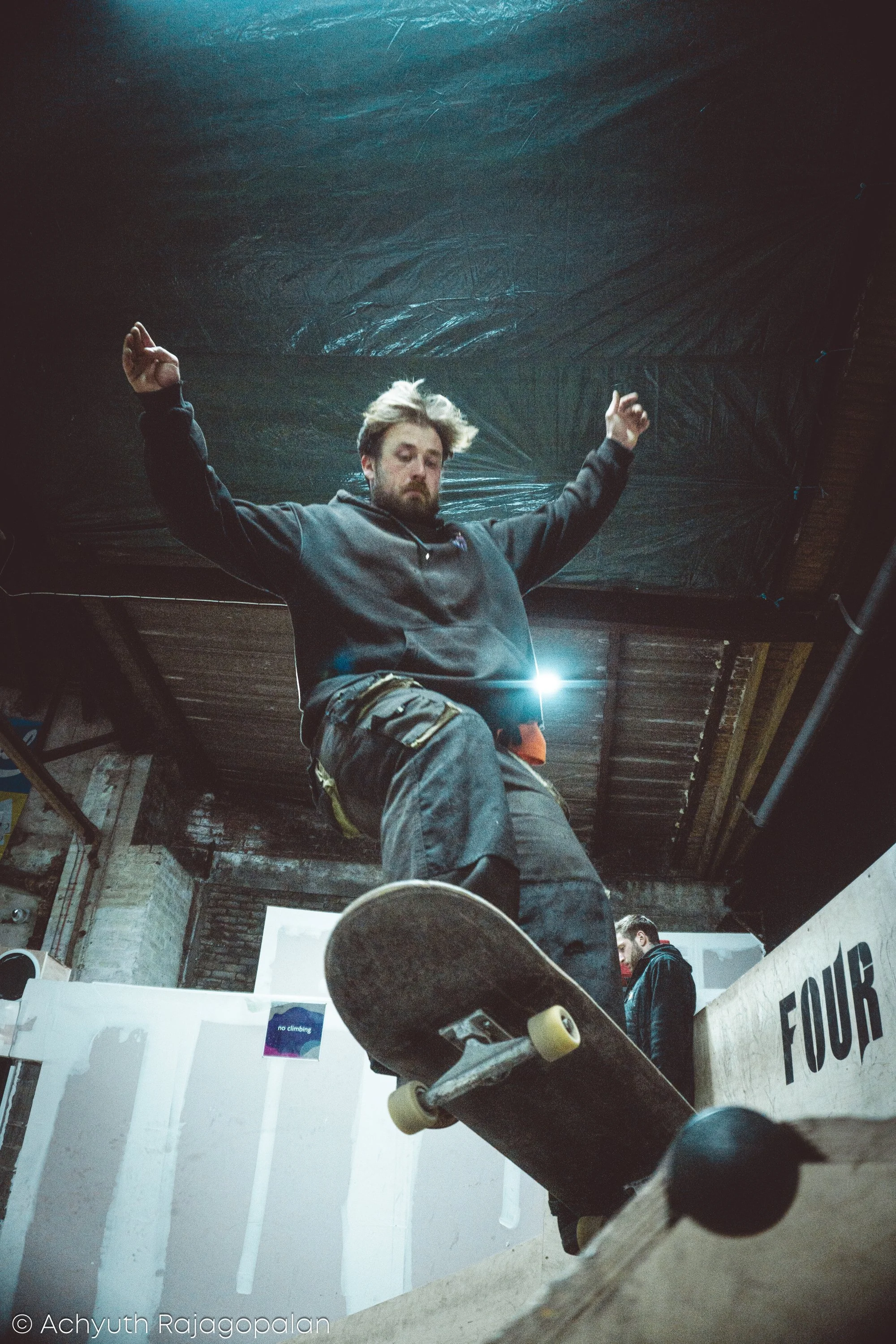 The image shows a young man with a beard, raising his arms in the air, standing in an indoor skateboarding area. The scene is from a low angle, with his skateboard visible in the foreground. The background includes a wall with a 'no climbing' sign an