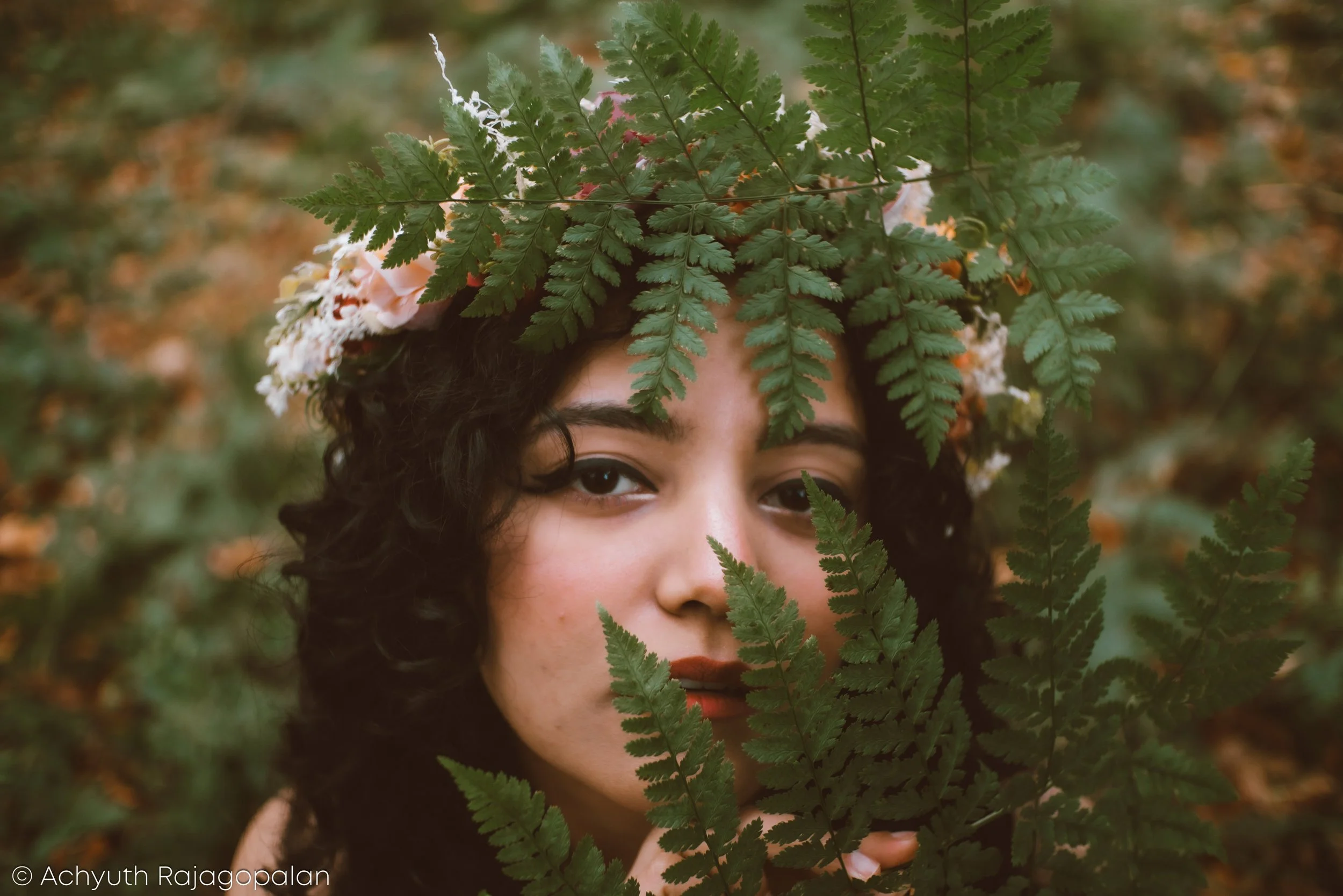 Person with dark curly hair partially covered by green fern leaves outdoors.