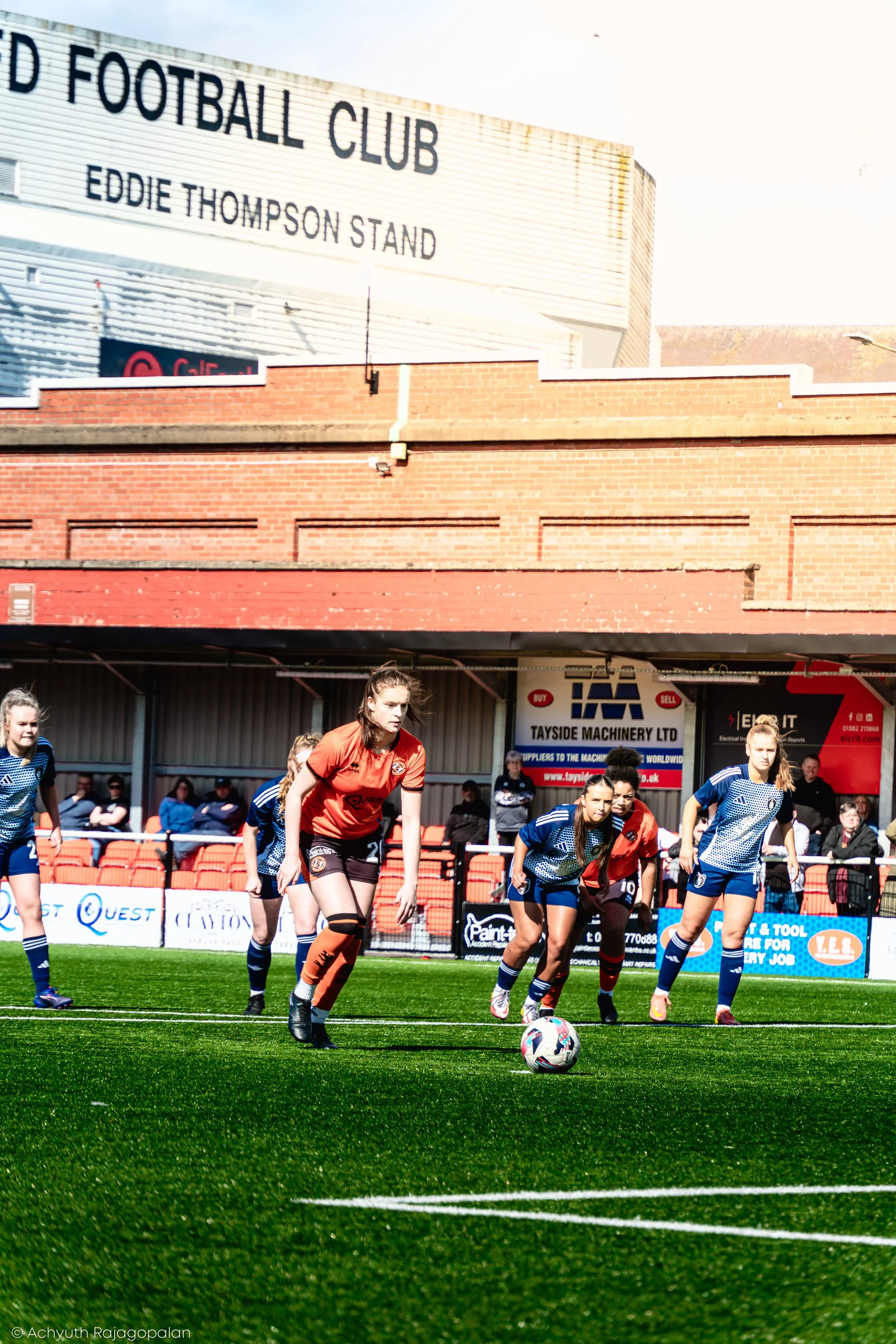 Women playing soccer on a green field during daytime with a group of spectators in the background and a building with a sign that reads 'Football Club' and 'Eddie Thompson Stand'.