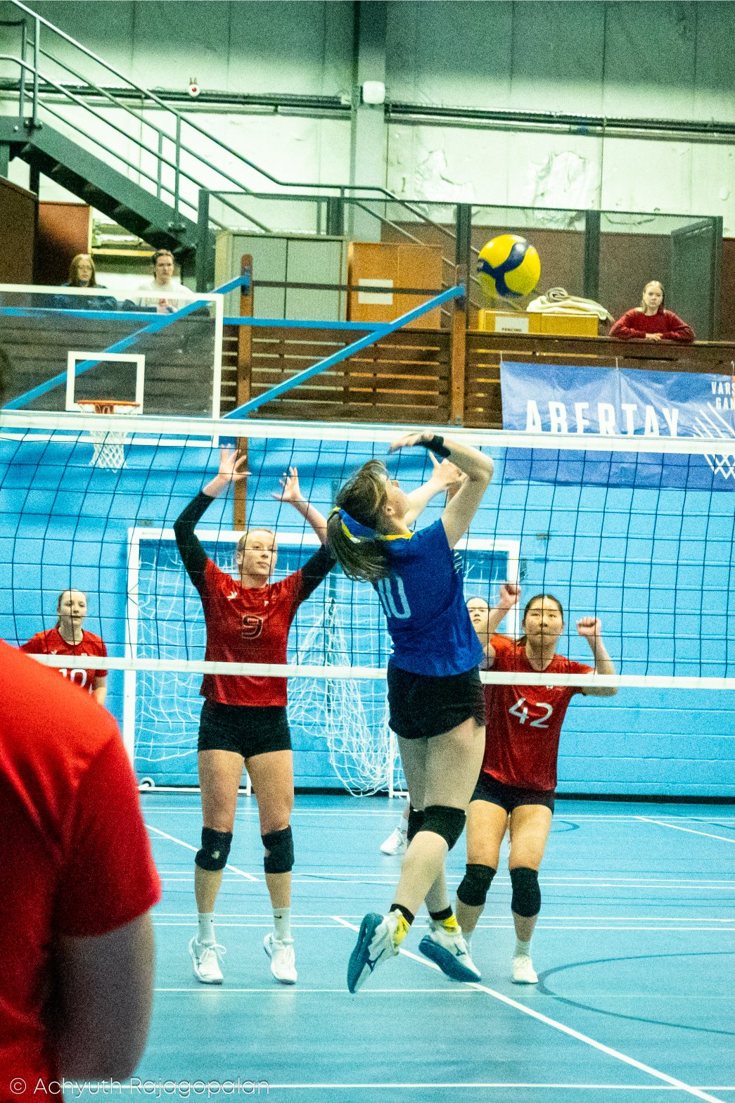 A volleyball match in progress with players on the court, one player in a blue uniform jumping to hit the ball, and three players in red uniforms preparing to block or receive, inside an indoor gymnasium.