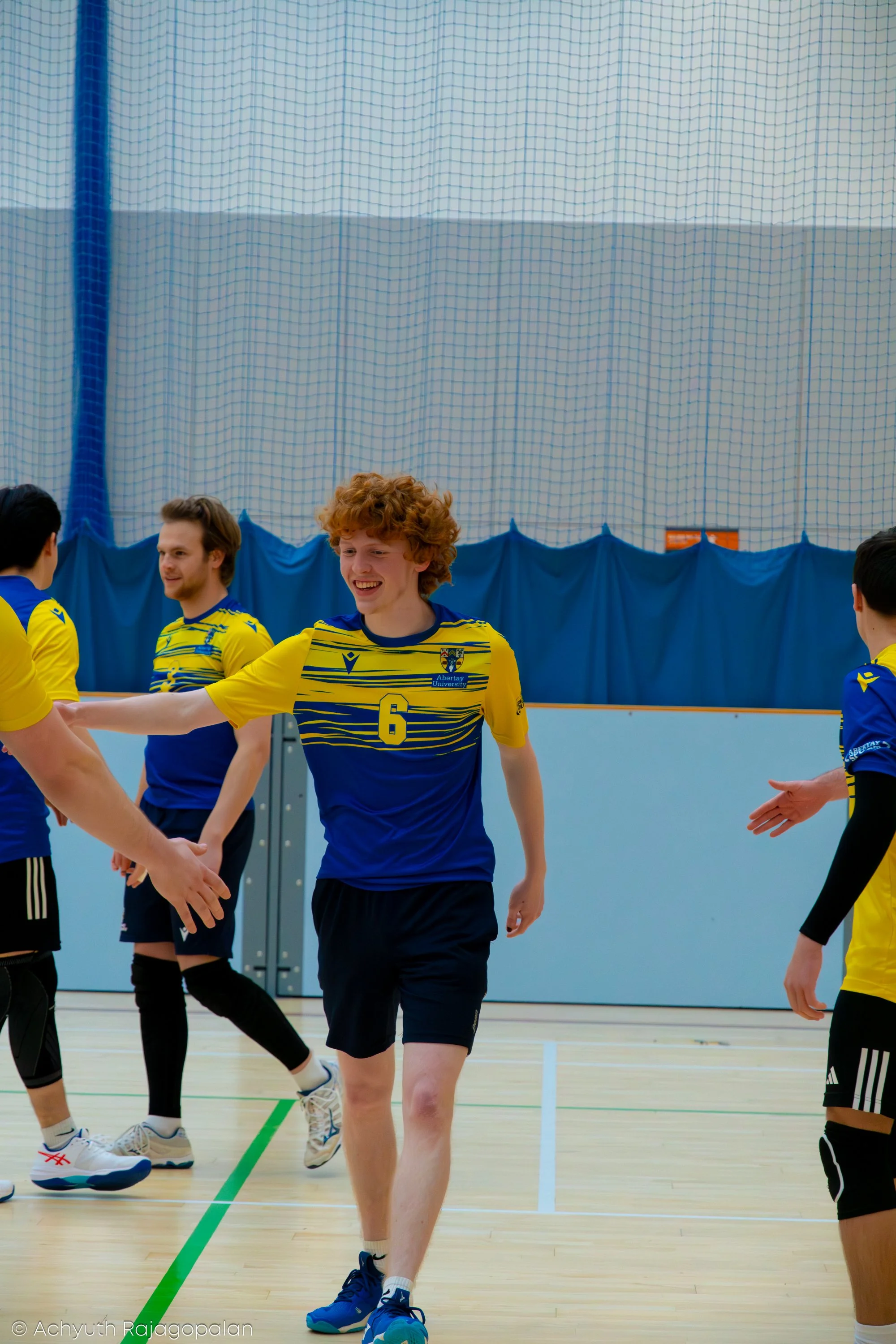 A group of young men in yellow and blue sports uniforms are standing and smiling inside a gymnasium. They appear to be volleyball players, engaged in a handshake or greeting after a game.