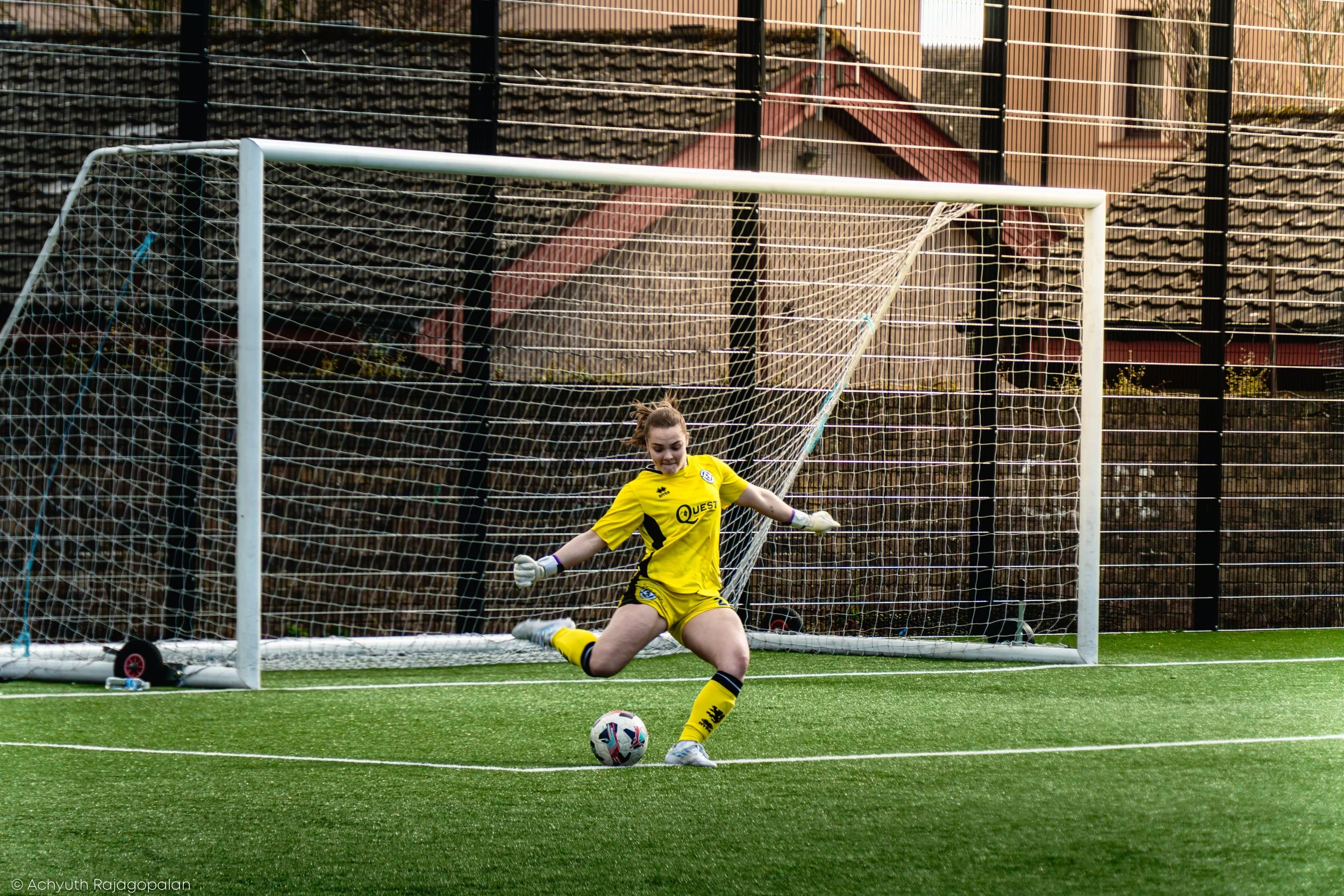 A female soccer goalkeeper in yellow uniform making a save in front of the goal, with a soccer ball on the ground nearby, on a green artificial turf field, with a fence and houses in the background.