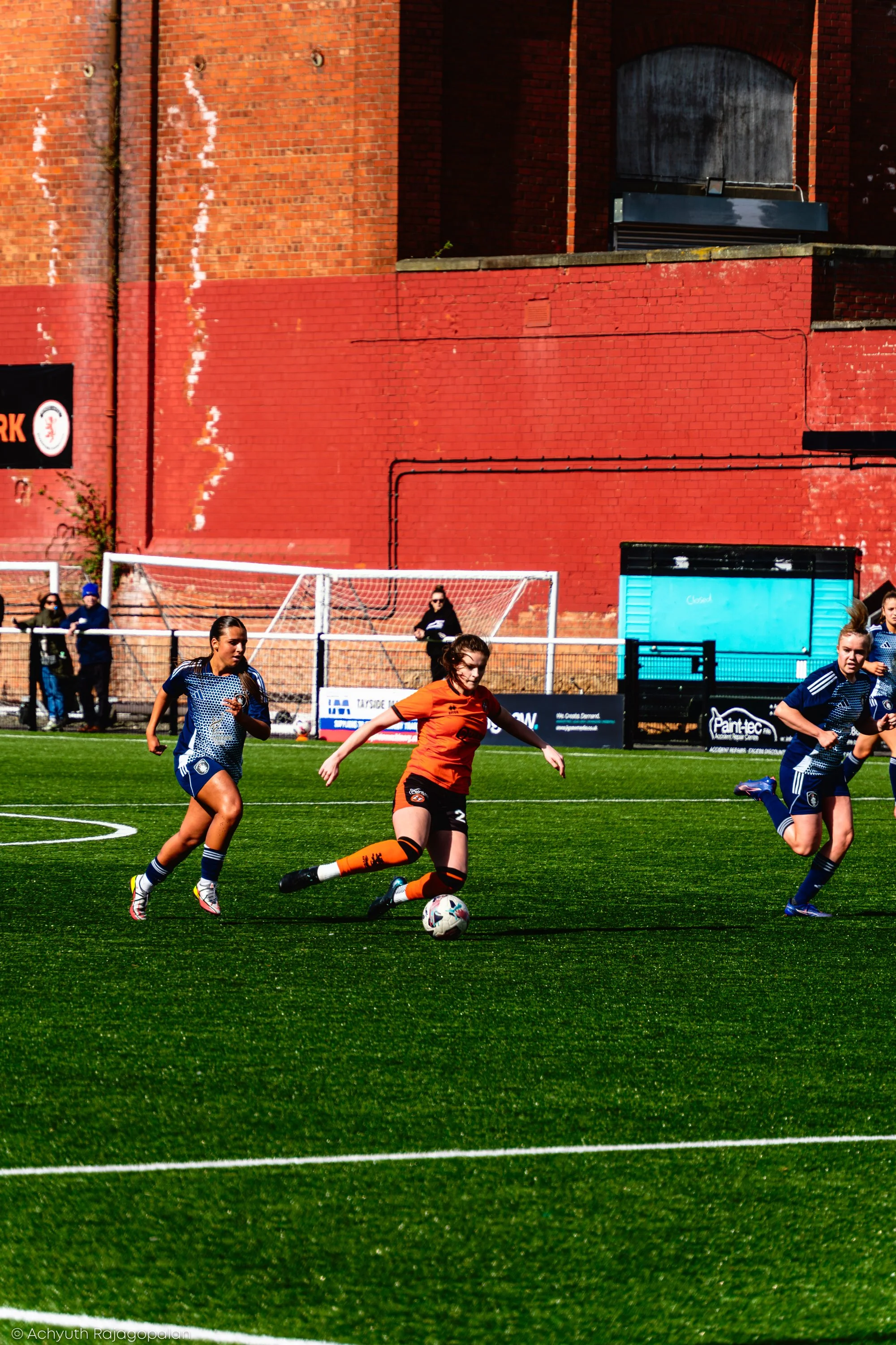 Women playing soccer on a field with a red brick wall in the background.