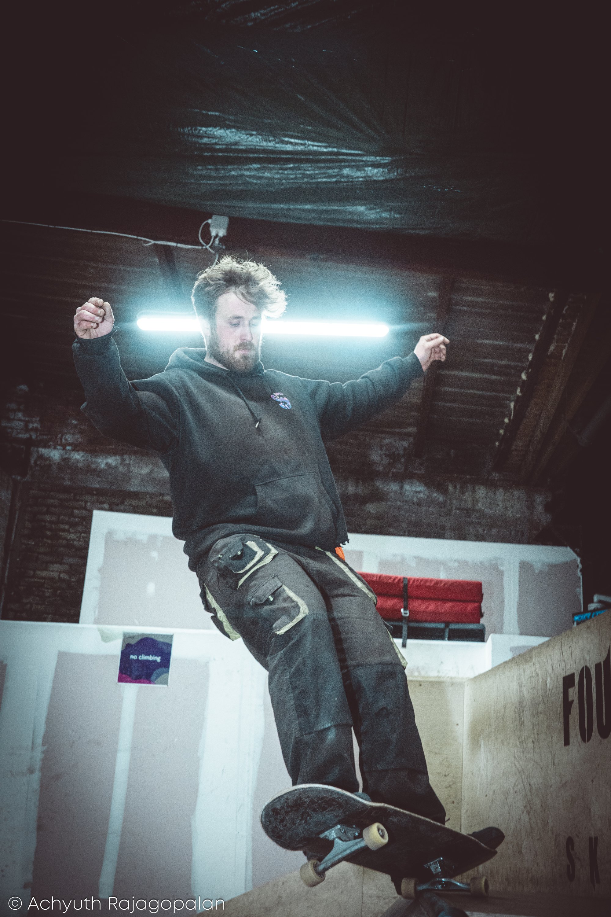 A man with a skateboard performing in an indoor skate park, with his arms raised and a focused expression on his face.