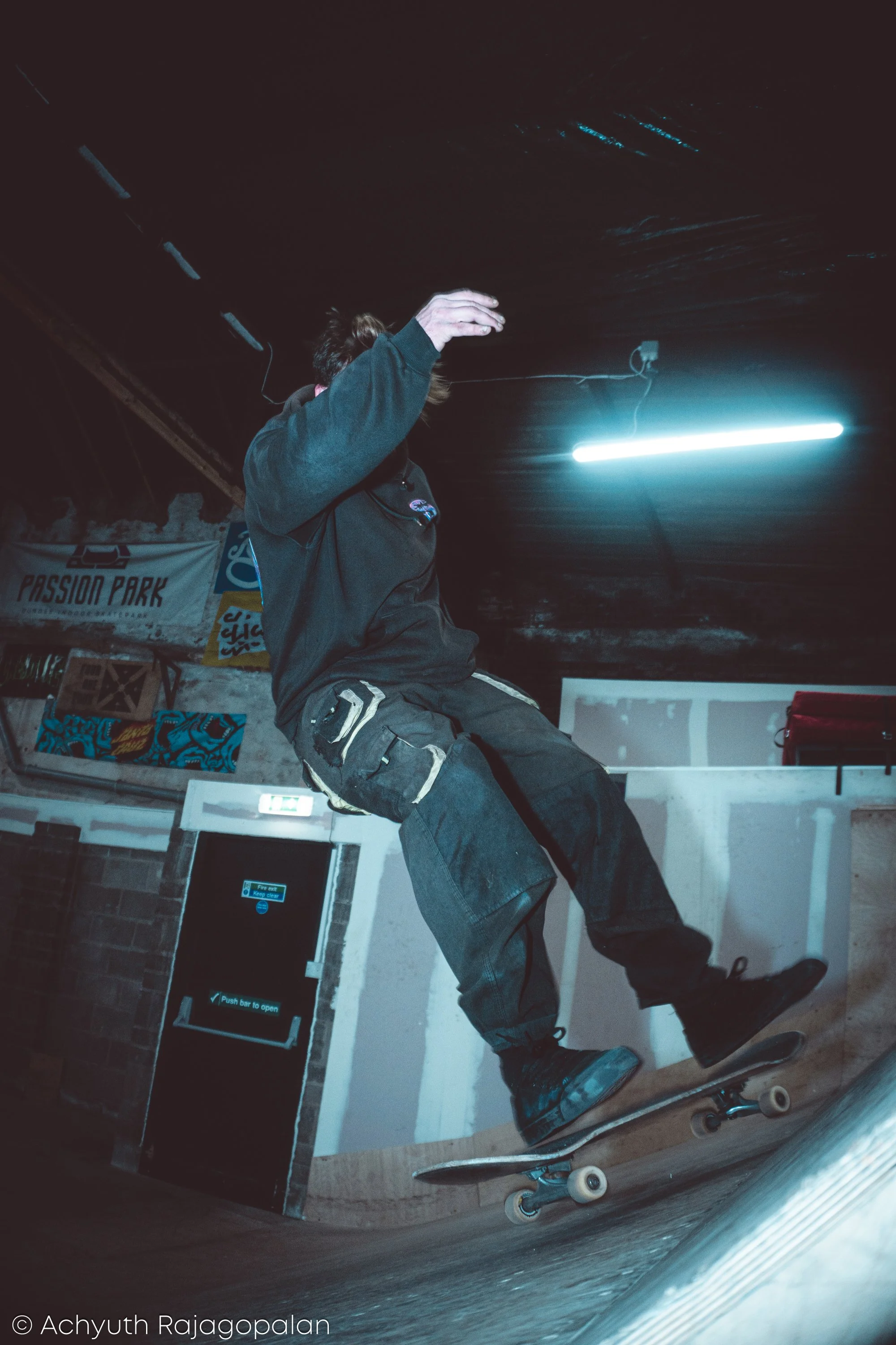 A person wearing a dark hoodie and cargo pants riding a skateboard indoors. The background has posters, a door with a sign, and a fluorescent light on the ceiling.