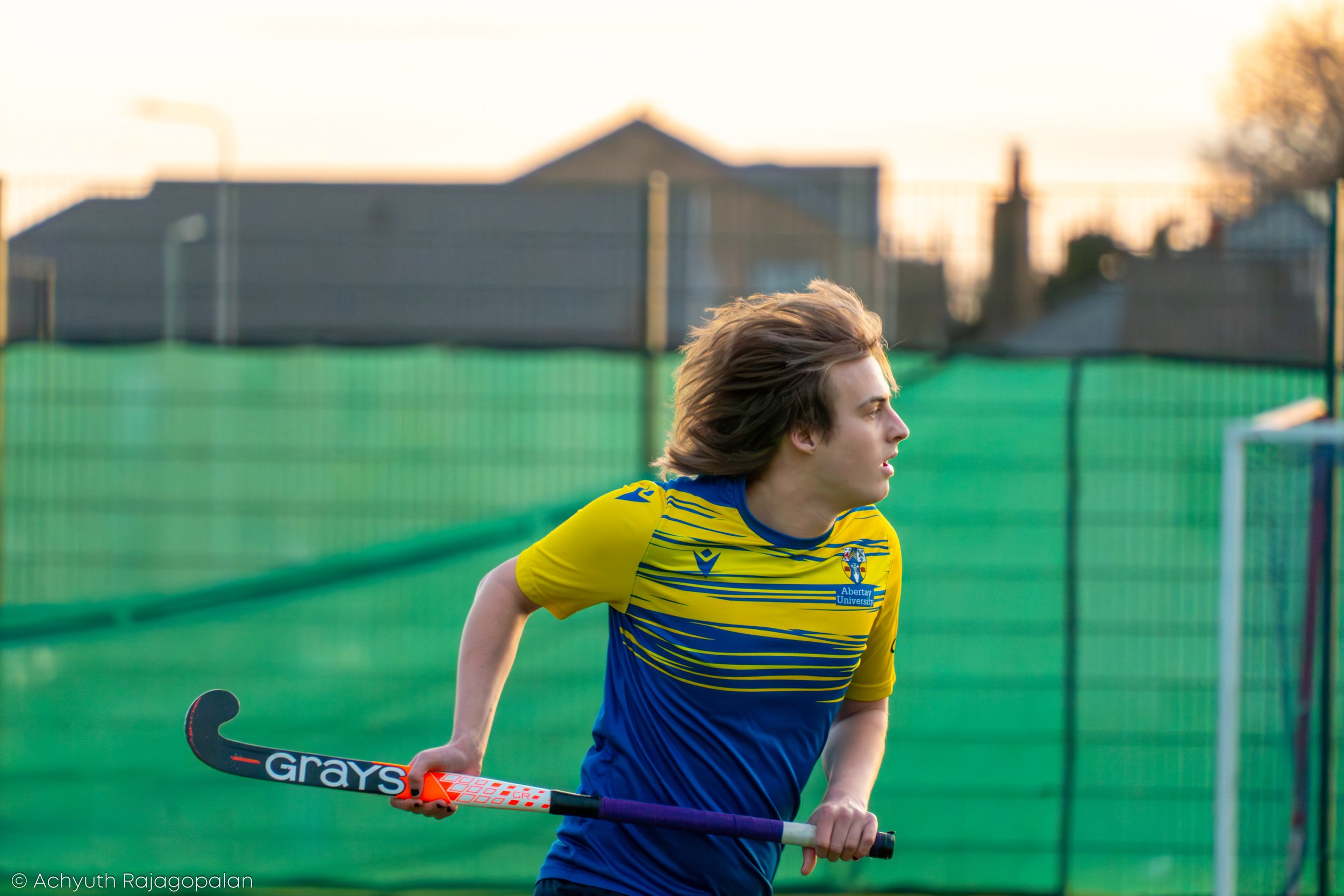 A young male field hockey player with long hair wearing a yellow and blue uniform, holding a hockey stick, on a sports field during sunset.