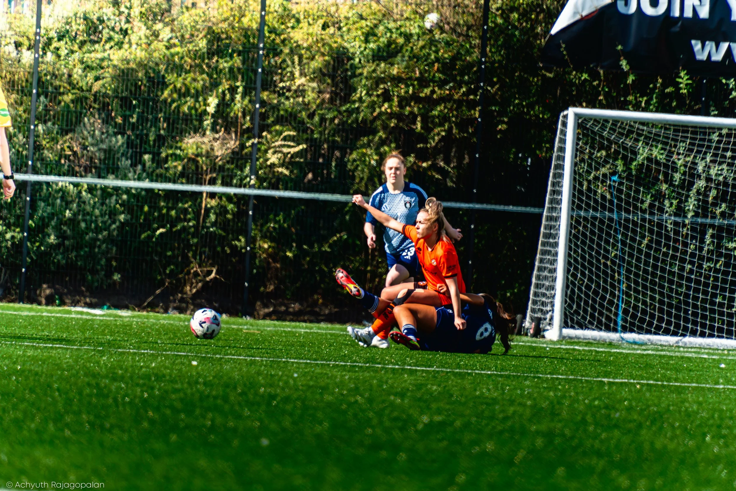 A soccer game with players near the goal, including a player in an orange jersey making a move to score and a goalkeeper in a black uniform trying to block the shot. The ball is on the grass near the goal line.