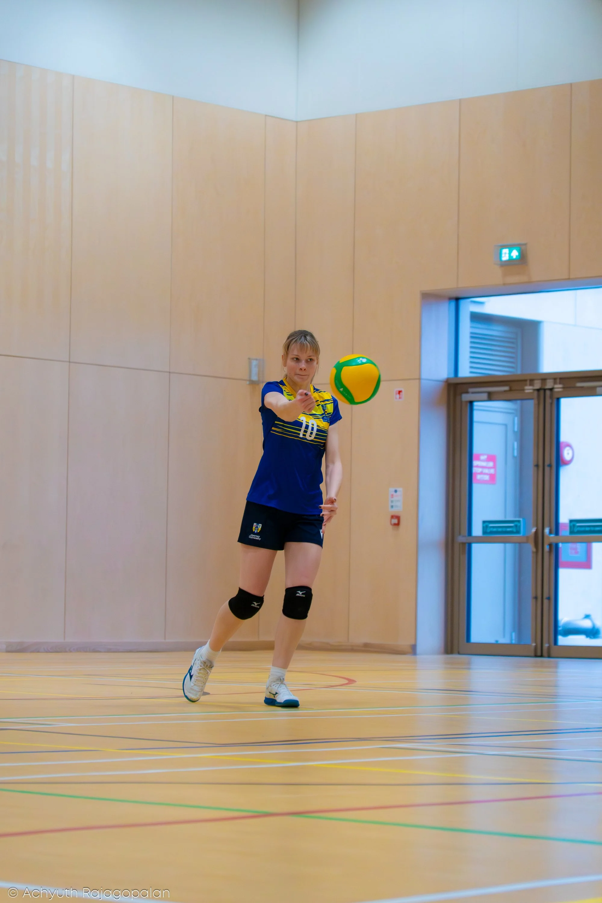 A female volleyball player in a blue and yellow jersey with the number 10, wearing knee pads, white sneakers, and black socks, is serving the ball in an indoor gymnasium with wooden walls and a yellow floor.