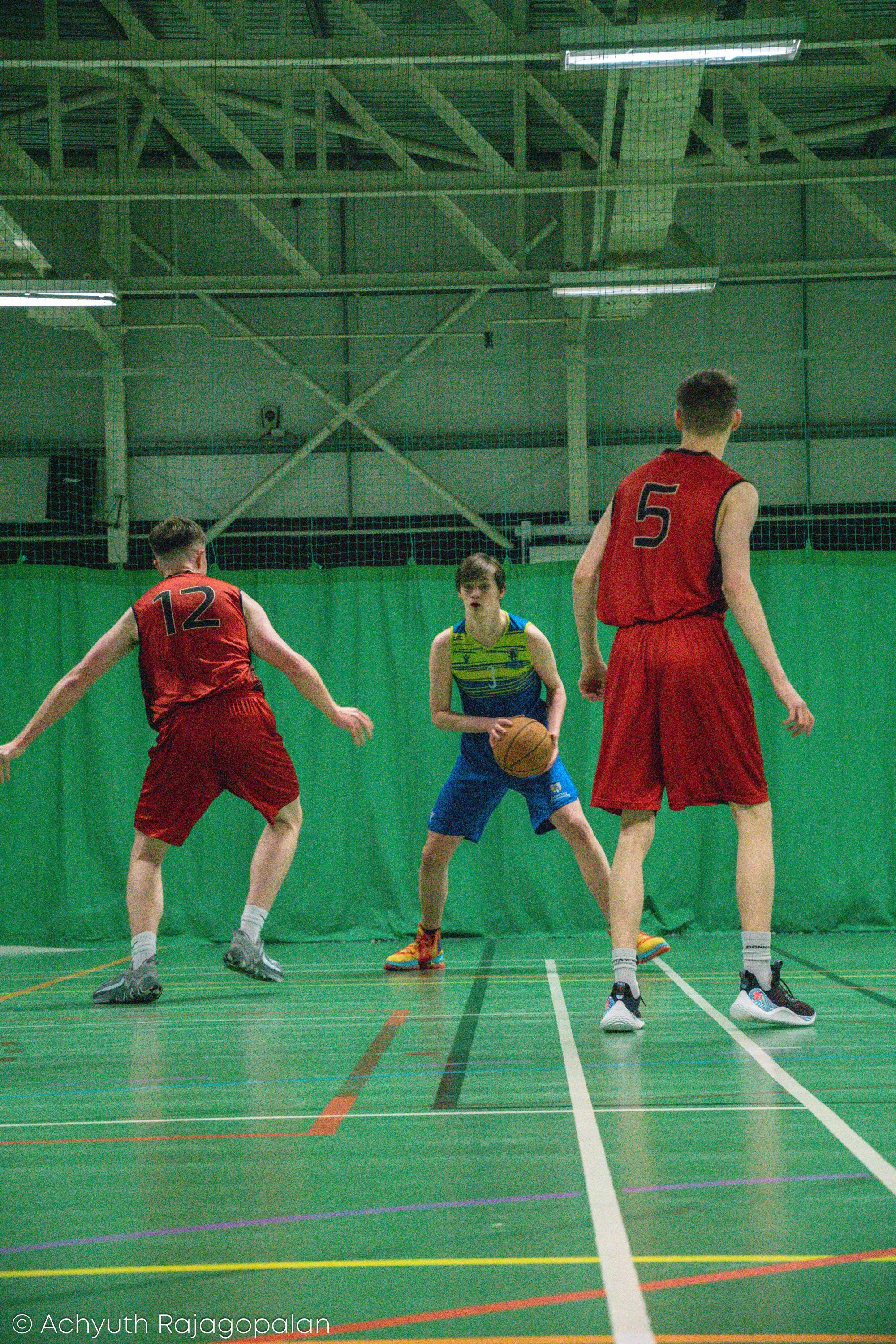 Three young males playing basketball on an indoor court with green walls and a high ceiling, two are wearing red jerseys with numbers 12 and 5, and one is in a blue and yellow jersey holding a basketball, seen from the back and front.