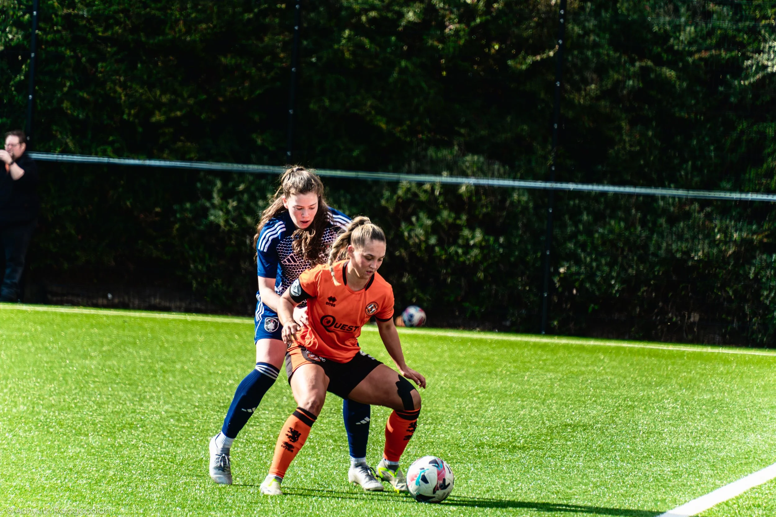 Two female soccer players competing for the ball during a match on a sunny day with green trees in the background.