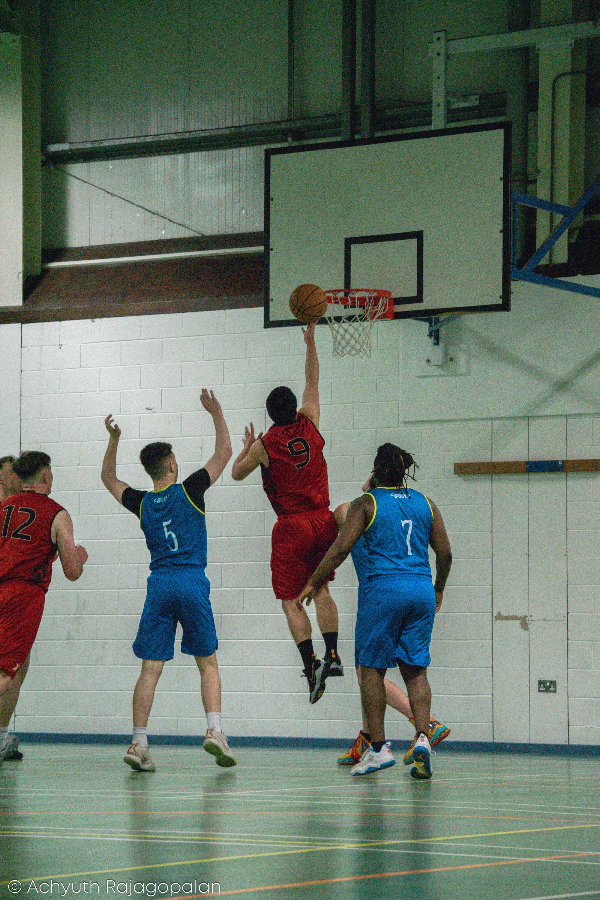 A basketball game taking place indoors with players reaching for the ball near the hoop. The player in red jersey number 9 is jumping to shoot or pass the ball, while players in blue jerseys attempt to block or defend. The hardwood court has markings