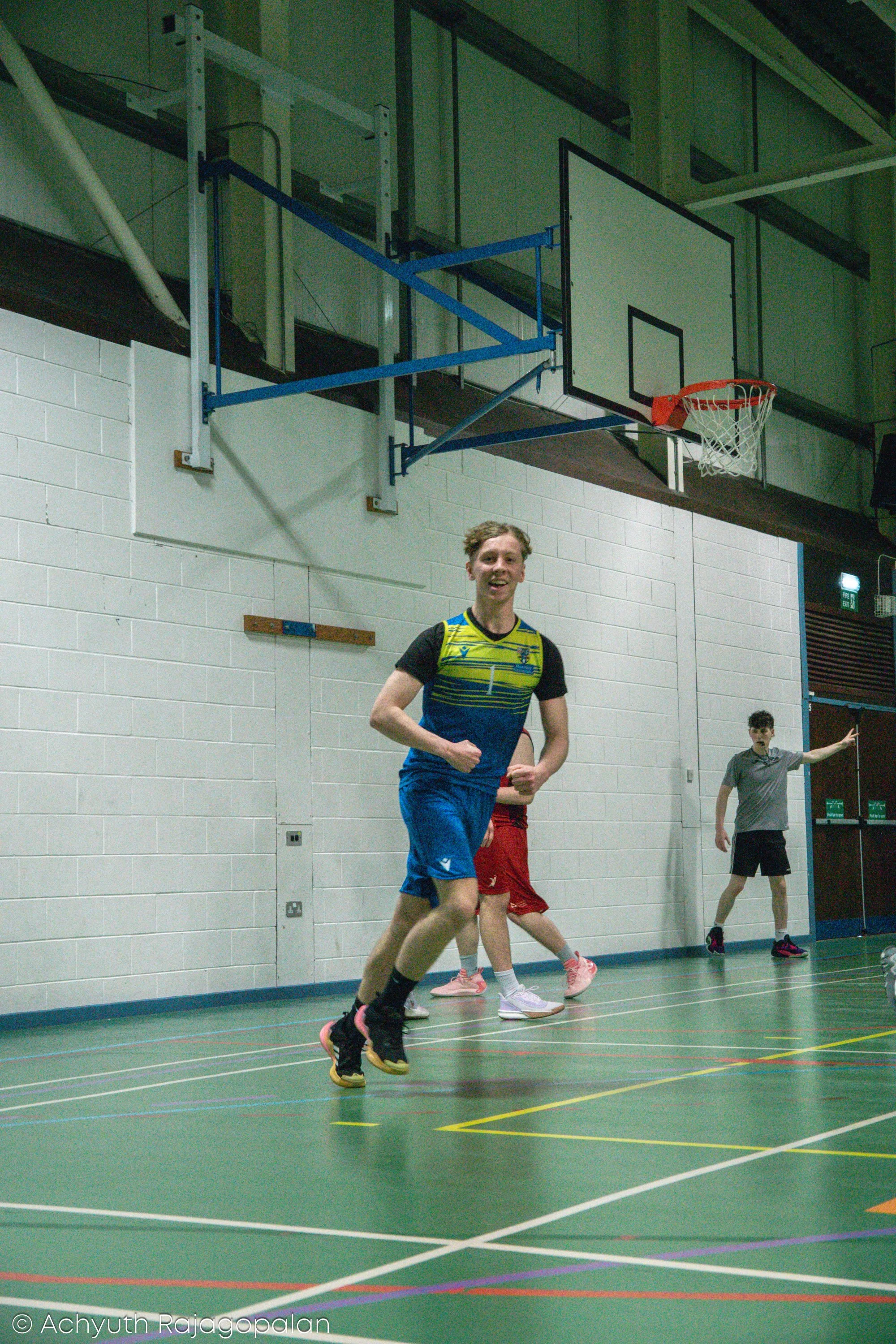 A young man running in an indoor gymnasium with a basketball hoop above. Other people are visible in the background, engaging in physical activity.