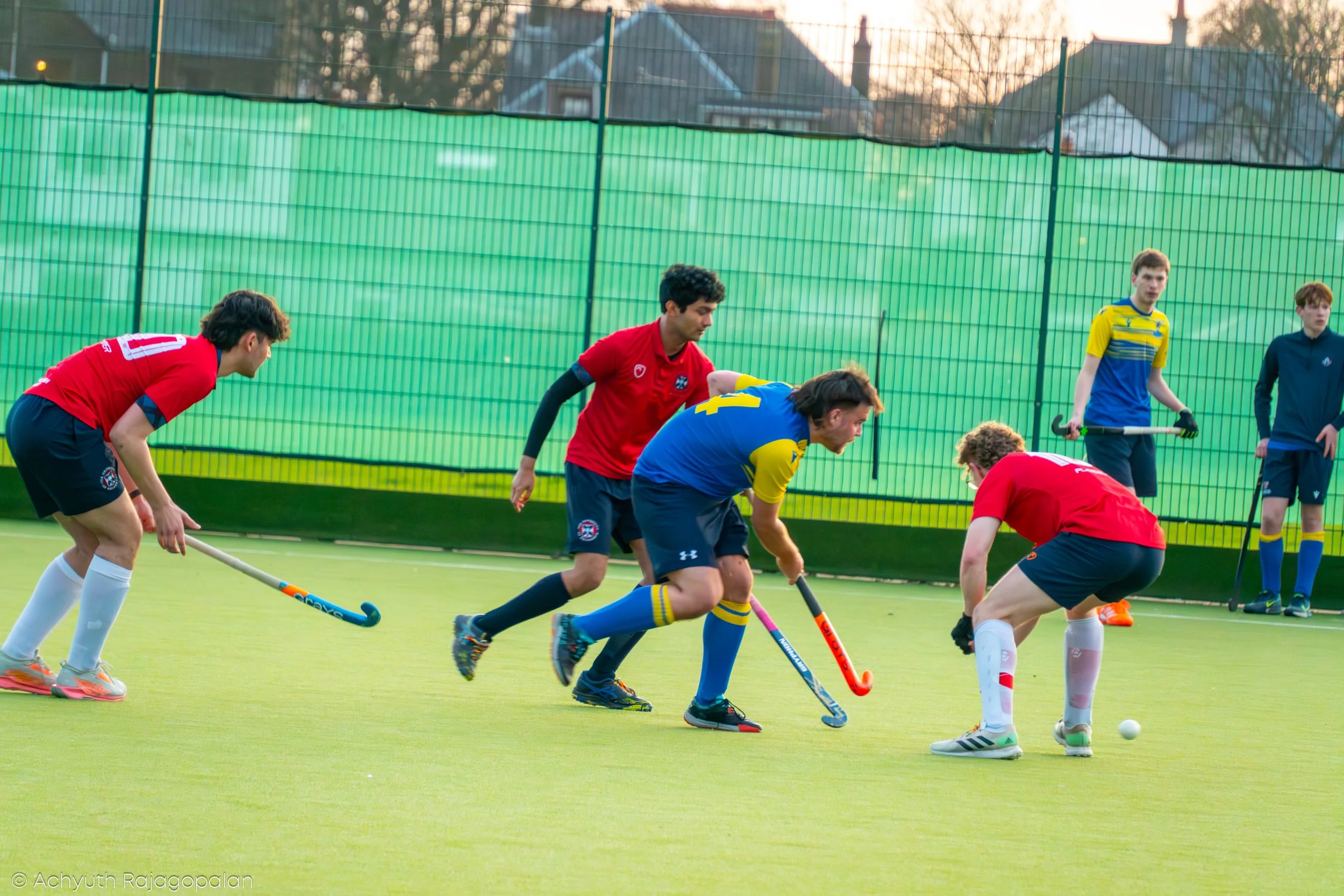 Group of young men playing field hockey on an outdoor green turf field enclosed by a tall green fence, with houses and trees in the background during sunset.
