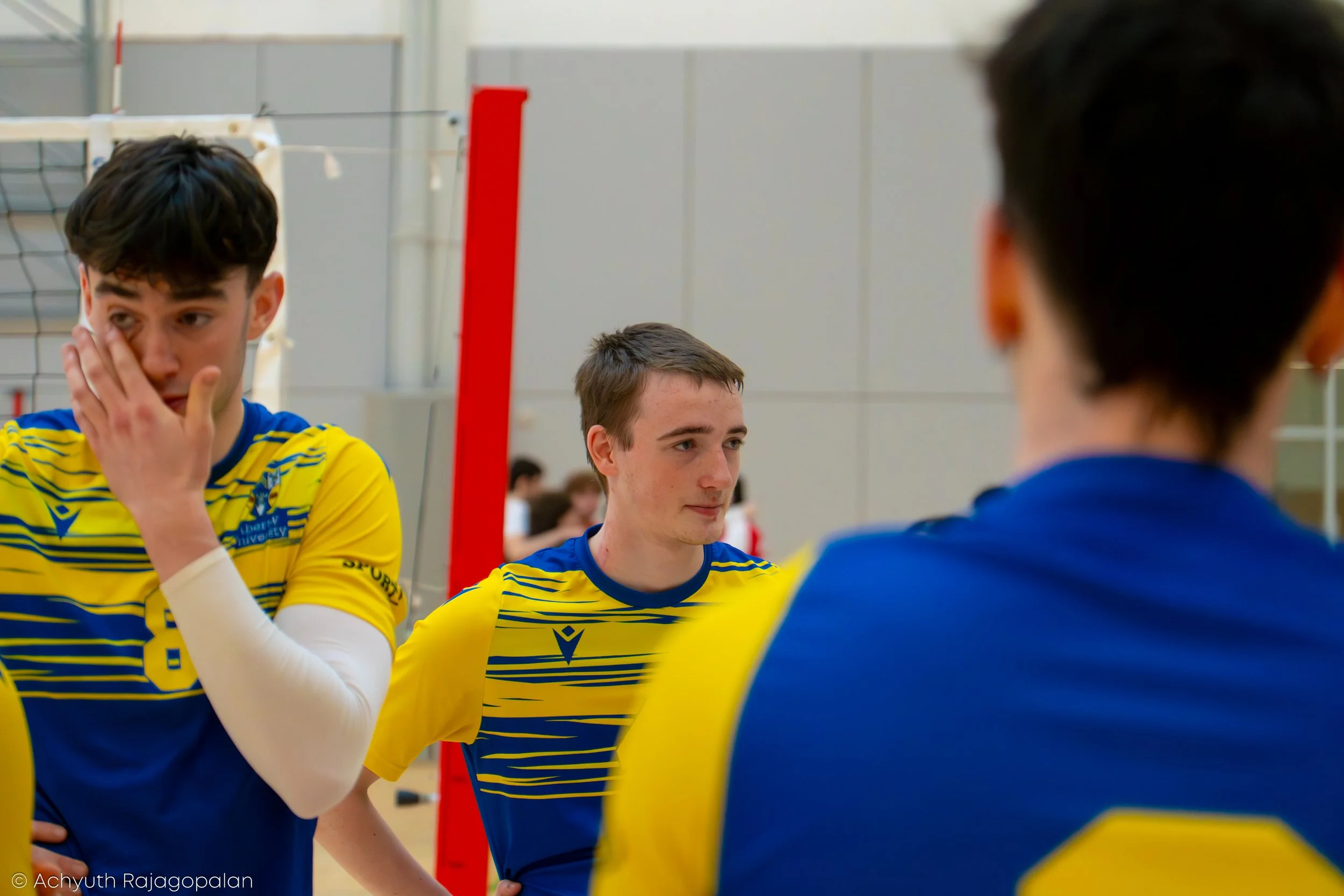 Three young male volleyball players in yellow and blue uniforms standing and talking in an indoor volleyball court.