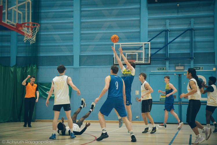 A basketball game with players in blue and white uniforms on an indoor court, with one player jumping to block a shot, others watching or moving, and a referee standing nearby.
