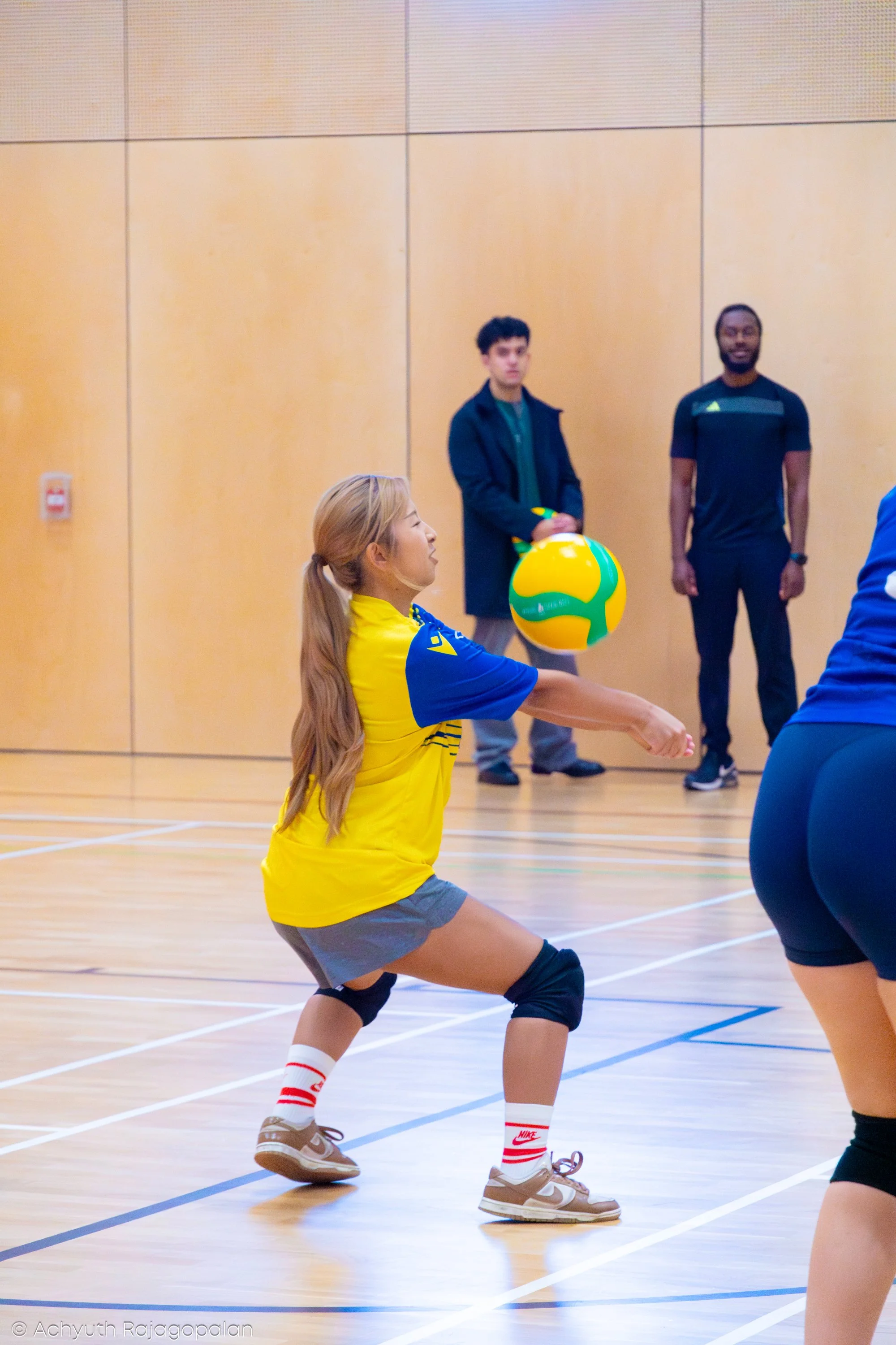 A woman in a yellow and blue sports outfit with knee braces playing volleyball indoors, preparing to bump the ball. Two men are in the background, standing against a wooden wall, watching her.
