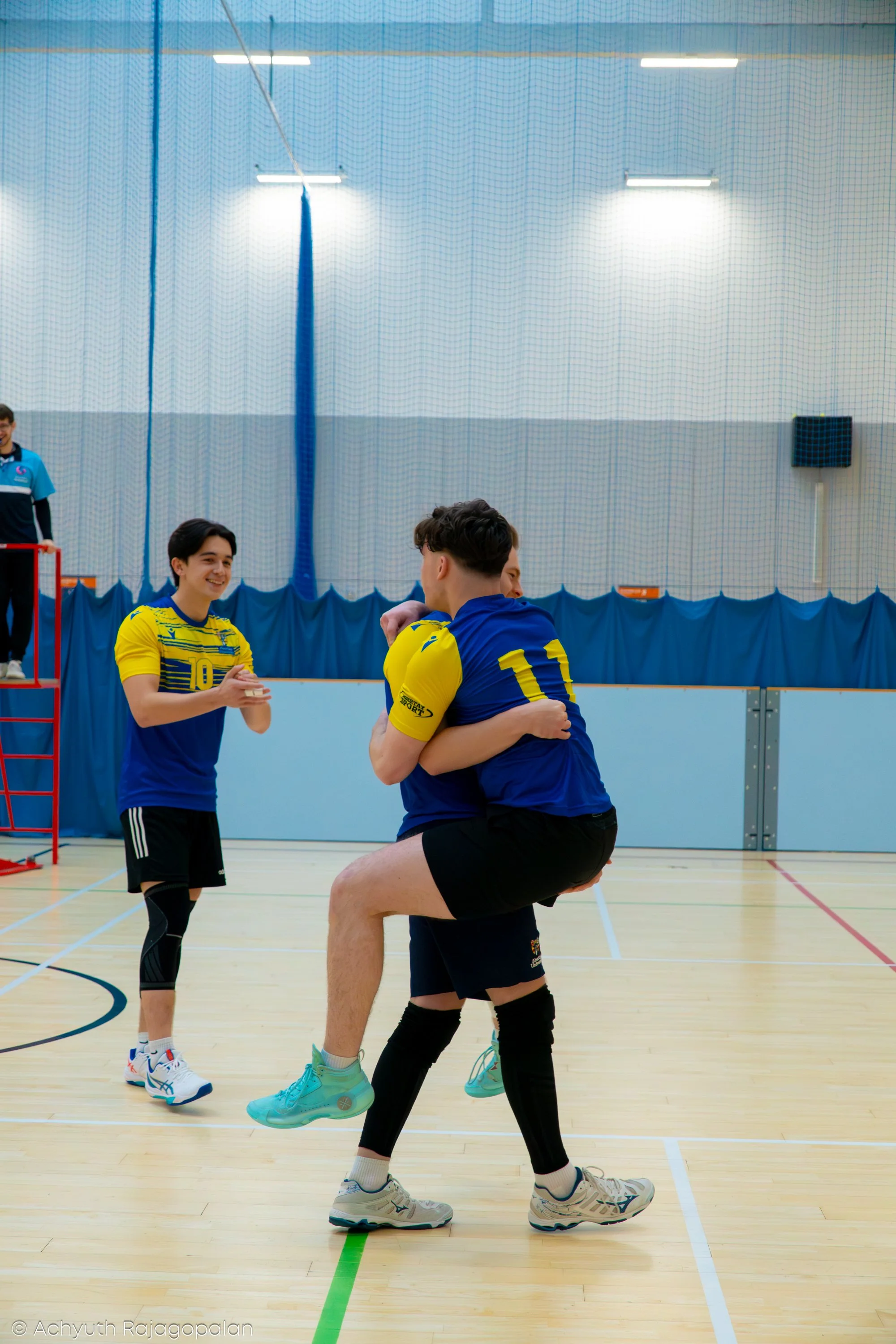 Two male volleyball players in blue and yellow jerseys celebrating on an indoor volleyball court, with a female referee standing in the background.