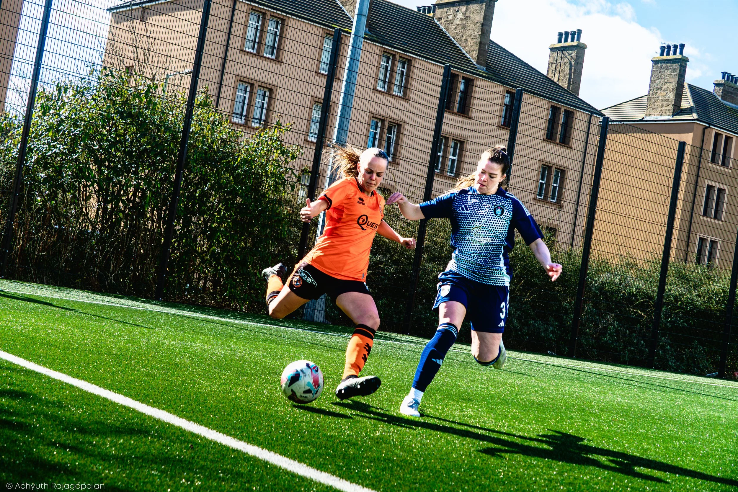 Two female soccer players competing for the ball on a green artificial turf field with residential buildings in the background, one wearing an orange jersey and the other wearing a blue jersey.
