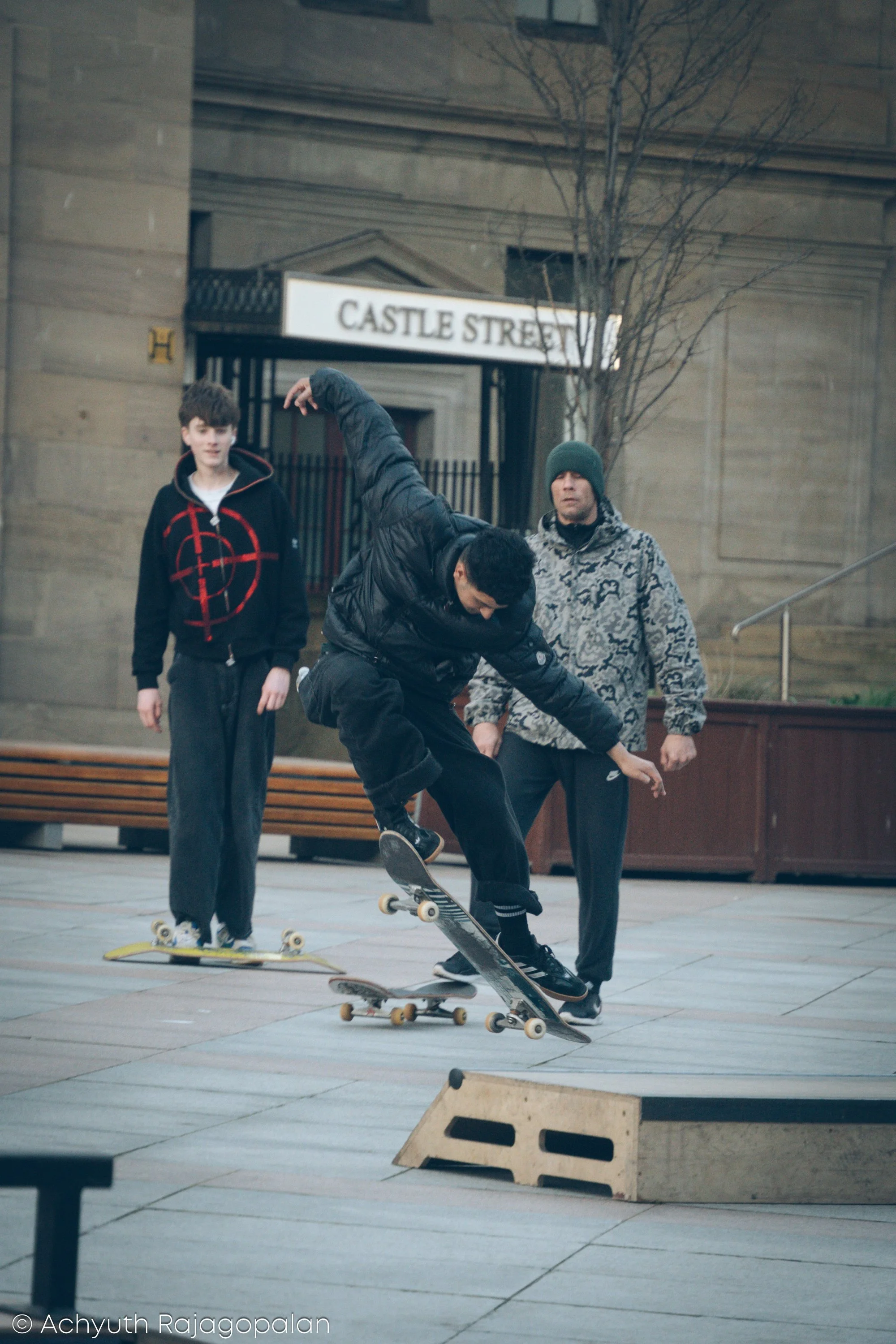 A group of young men skateboarding outdoors in an urban area. One skater is performing a trick, while others watch, with a building and street sign that reads 'Castle Street' in the background.
