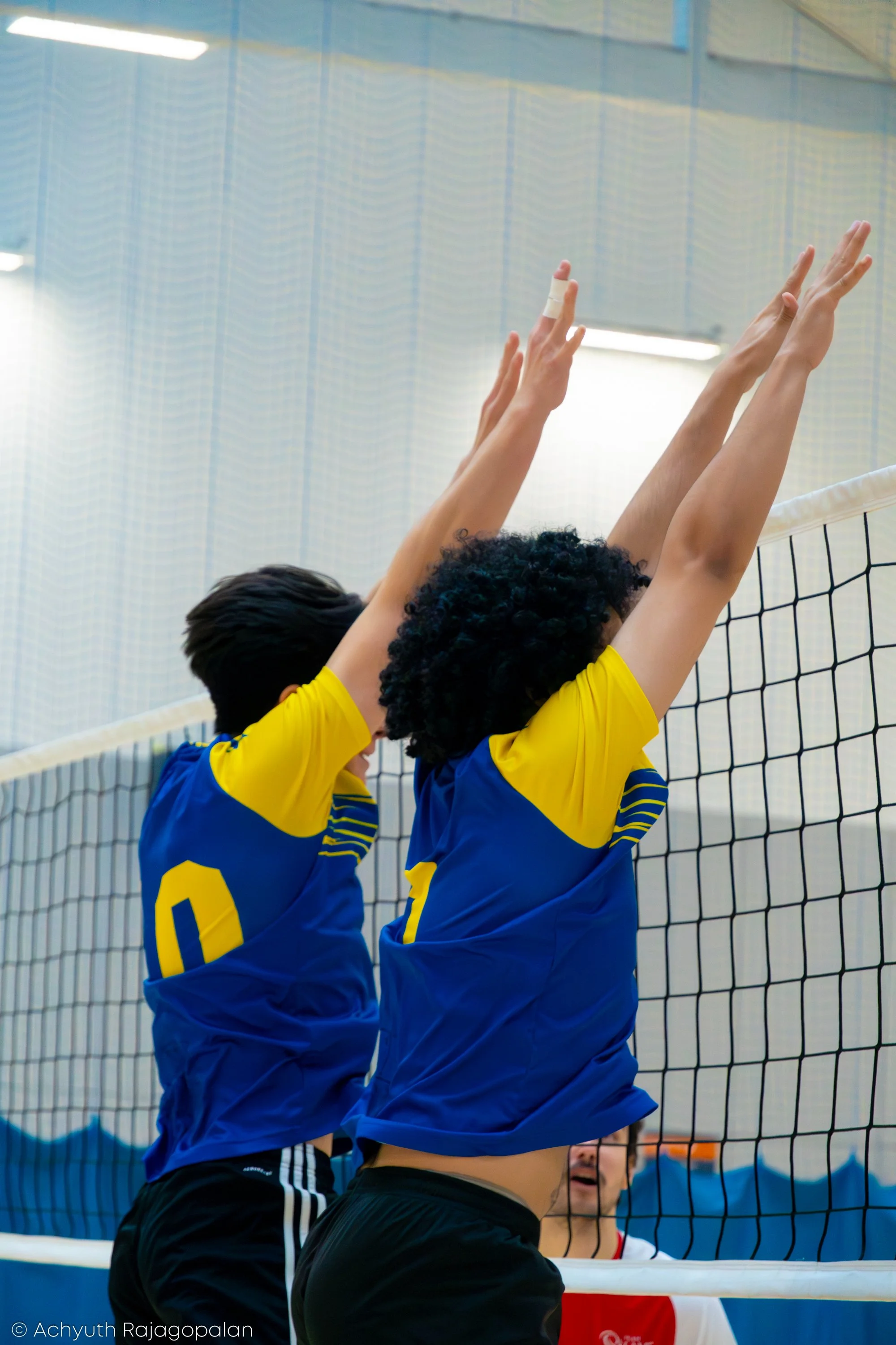 Two volleyball players in blue and yellow jerseys jump to block a ball at the net, with a volleyball in midair. Another player with a white and red jersey is visible beneath the net. The indoor court has a blue wall and a light-colored ceiling.