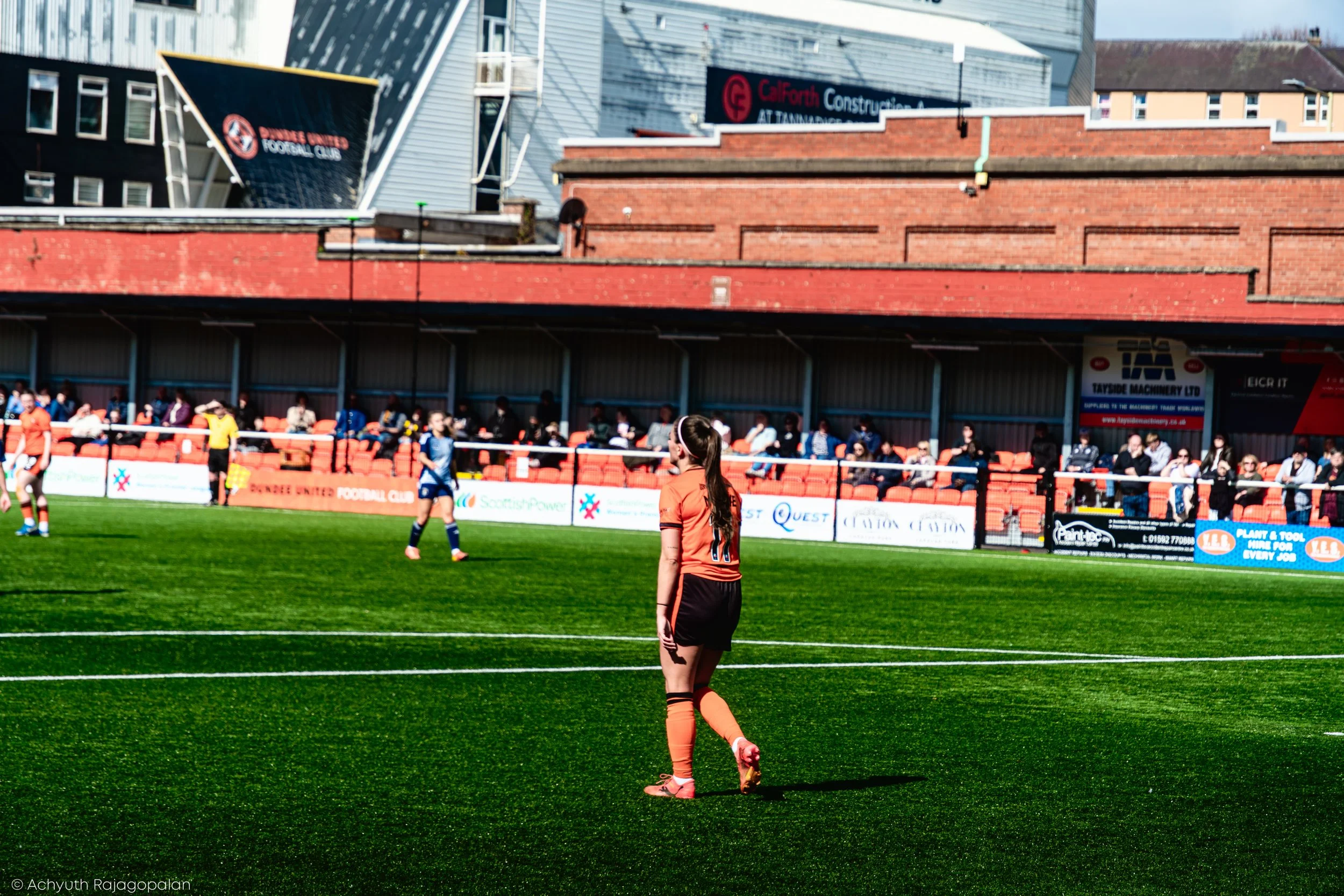A soccer match is taking place on a green field at a stadium with spectators sitting in the stands. Several players are visible, with a girl in an orange jersey and black shorts walking on the field. In the background, there are advertisements and ba