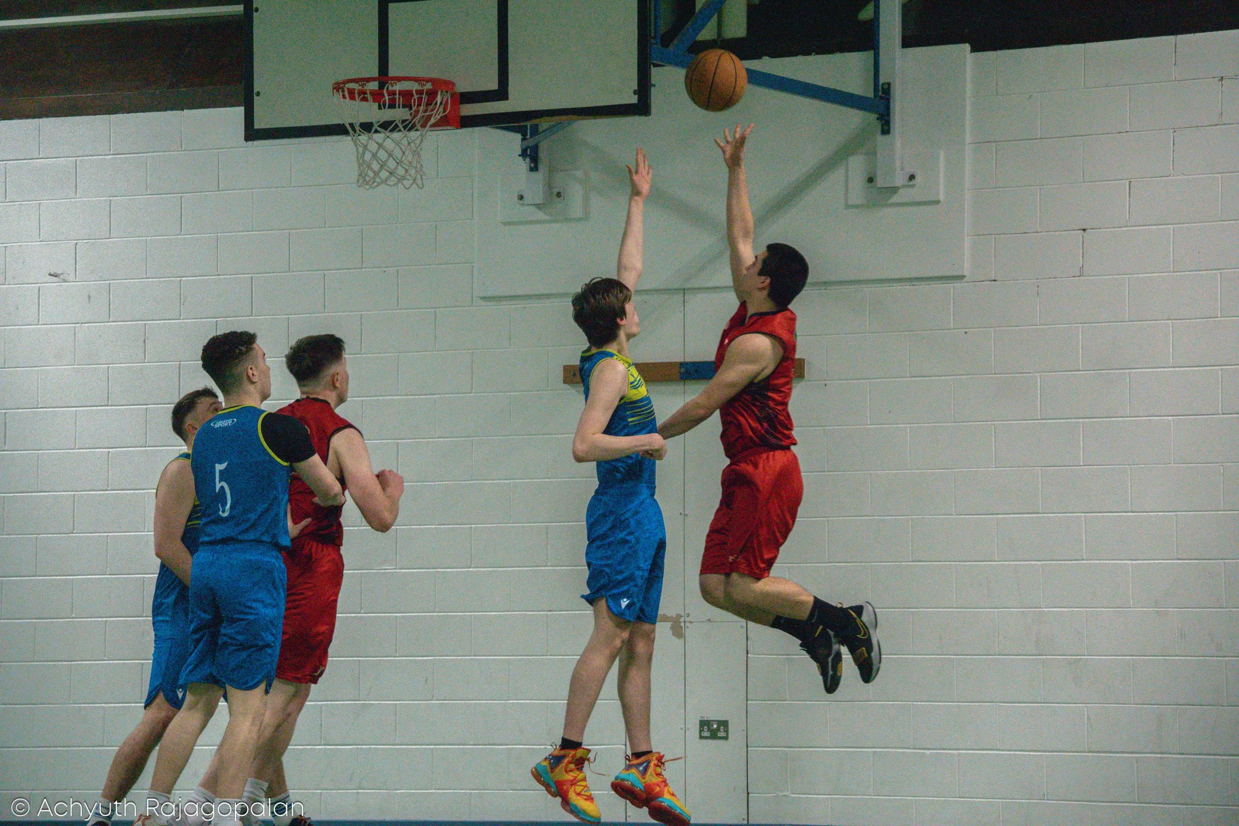 Two basketball players are jumping for a rebound near the hoop while three other players watch. The player in red is jumping to block or contest the shot made by a player in blue.