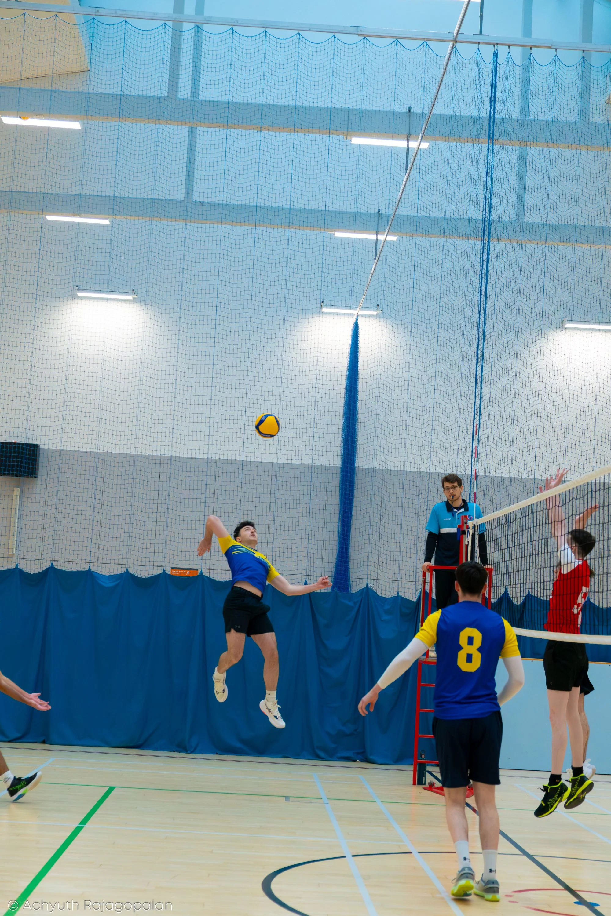 Indoor volleyball game with players jumping to hit the volleyball near the net. One player in yellow and blue jersey jumps, another player in red attempts to block the ball, referee watching the game.