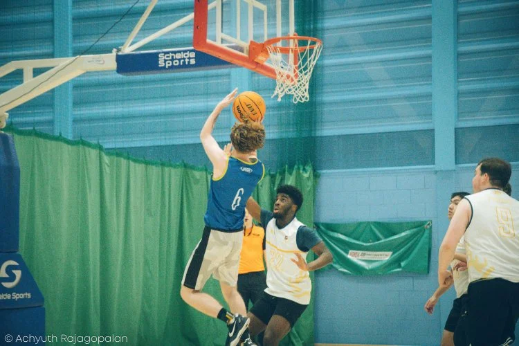 Basketball player in blue jersey jumping to make a shot at the hoop during a game, with opponents in white jerseys watching.