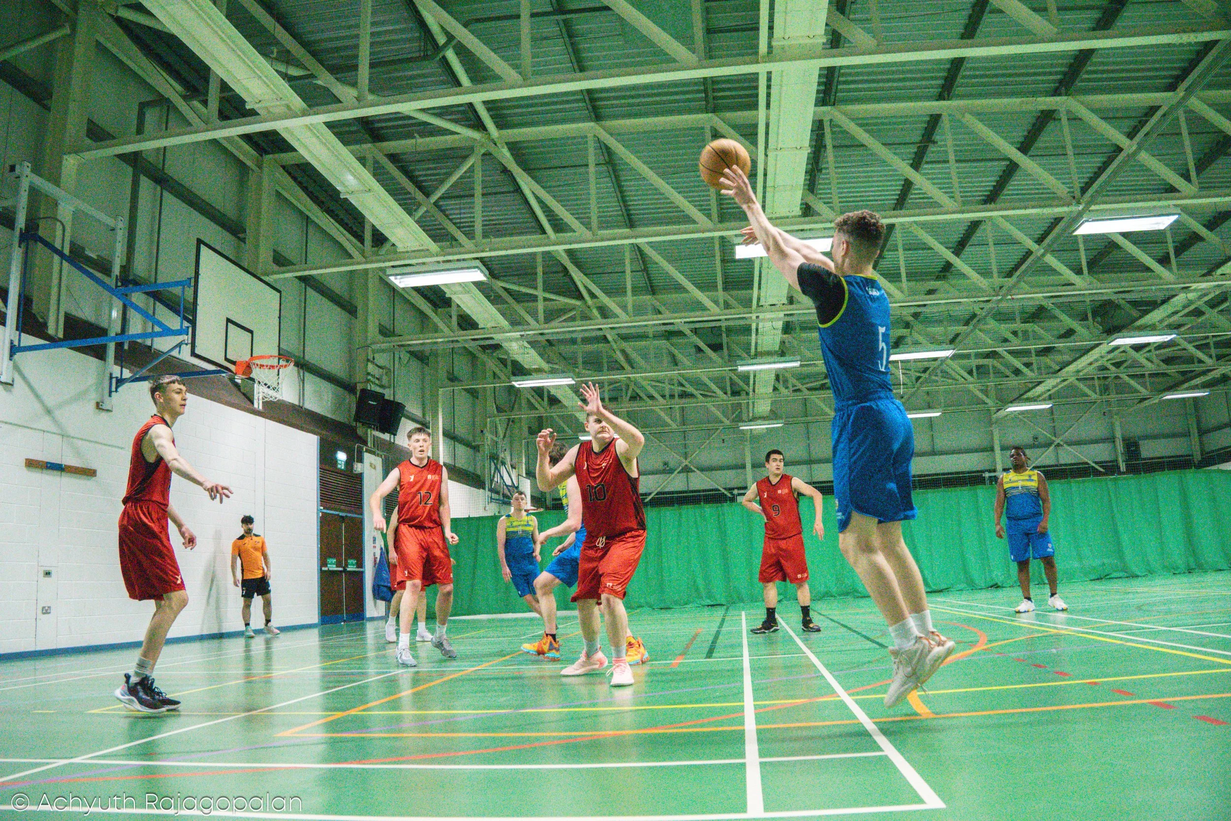 A group of young men playing basketball indoors on a green court. One player in blue jumps to take a shot while others in red are defending. The gym has a high ceiling with metal trusses and basketball hoops.