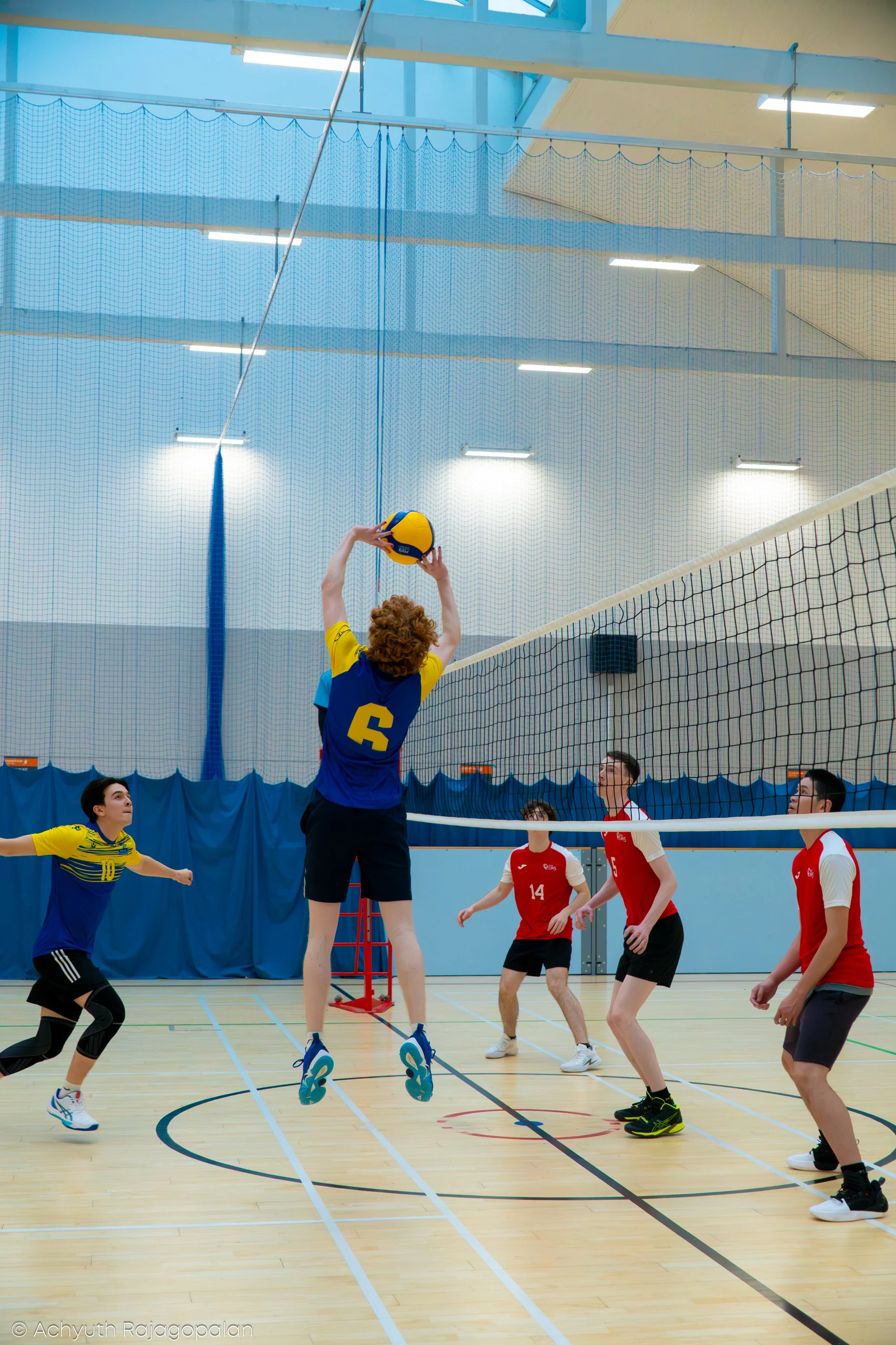 A volleyball game in progress at an indoor sports facility. A player with curly hair in a yellow and blue jersey, number 6, jumps to hit the ball, while a teammate with a yellow and blue jersey, number 10, prepares to assist. The opposing team in red